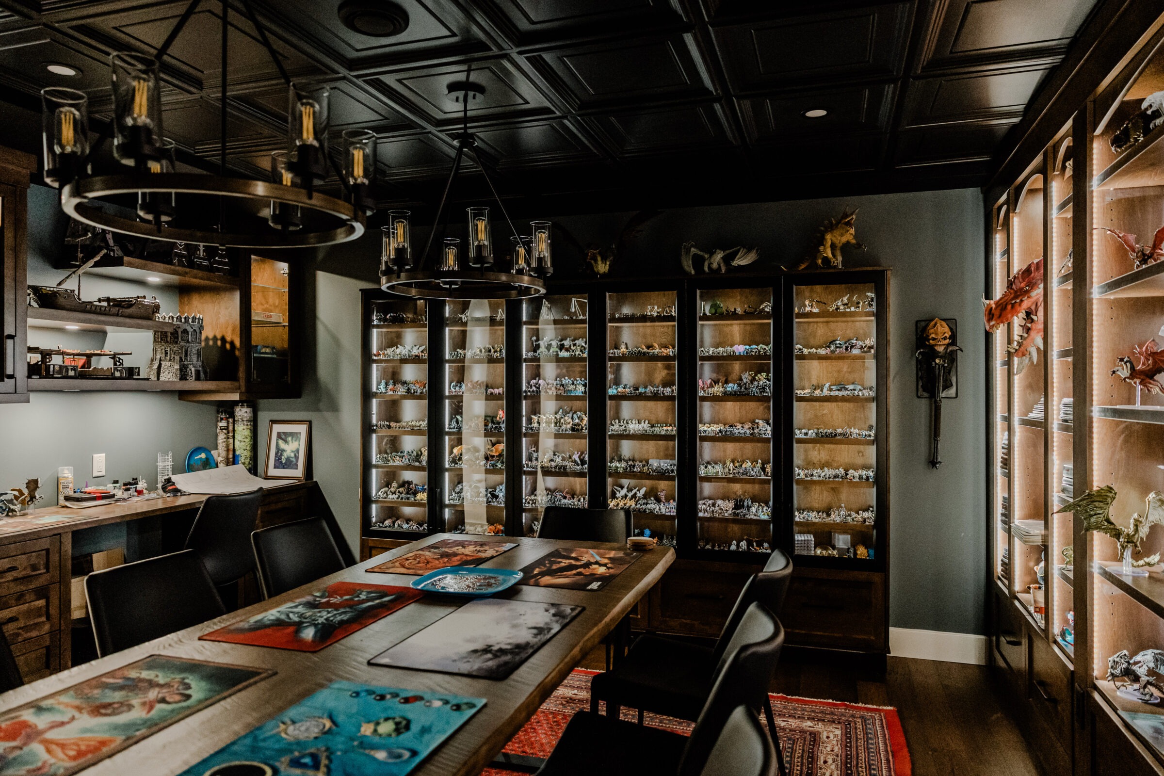 A dimly lit room featuring an extensive collection of miniatures on shelves, a wooden table with colorful mats, and vintage-style chandeliers.