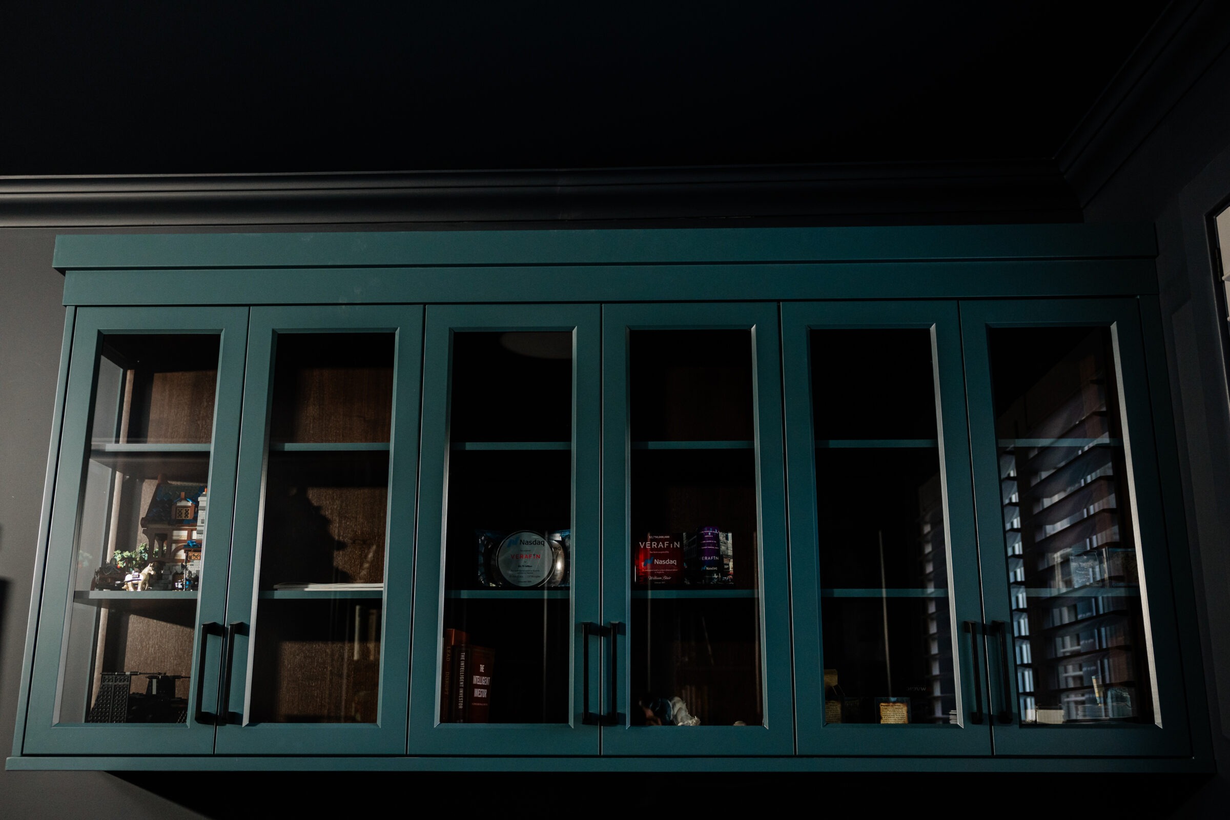A teal-colored, glass-paneled cabinet with various items on shelves, including books and decor, against a dark wall in dim lighting.