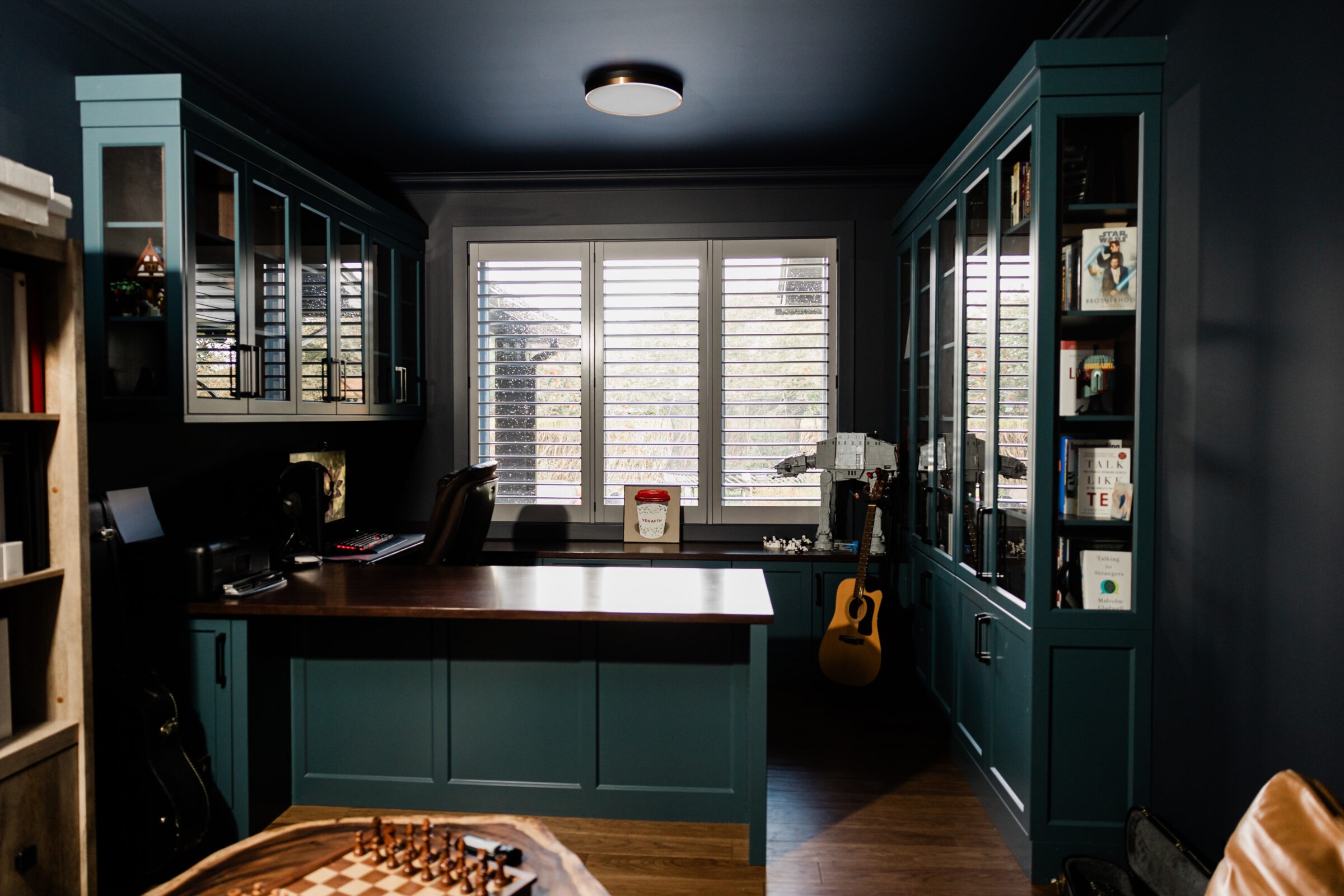 A cozy home office with dark blue walls, wooden floors, a desk with a computer, bookshelves, a guitar, and shutters partially open on the windows.