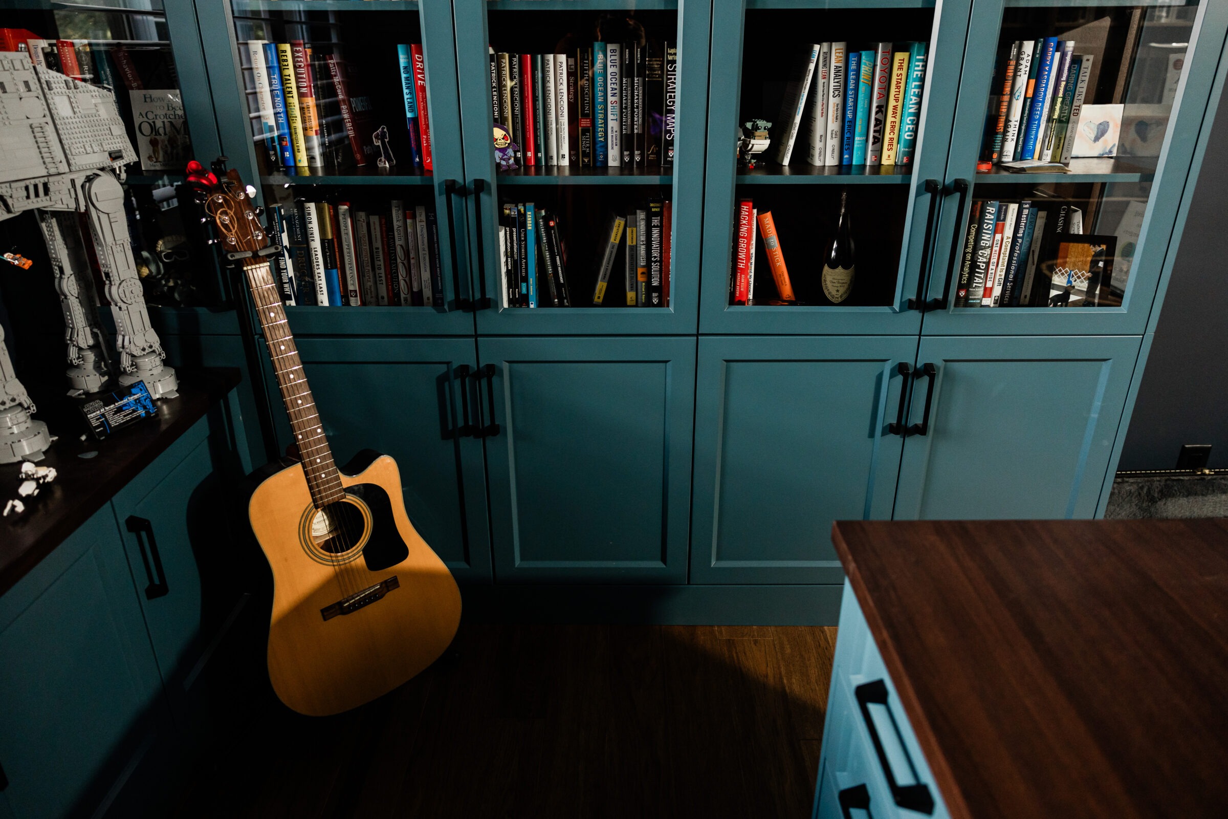 A cozy room featuring a large turquoise bookshelf filled with books, a guitar leaning against it, Star Wars LEGO models, and hardwood flooring.