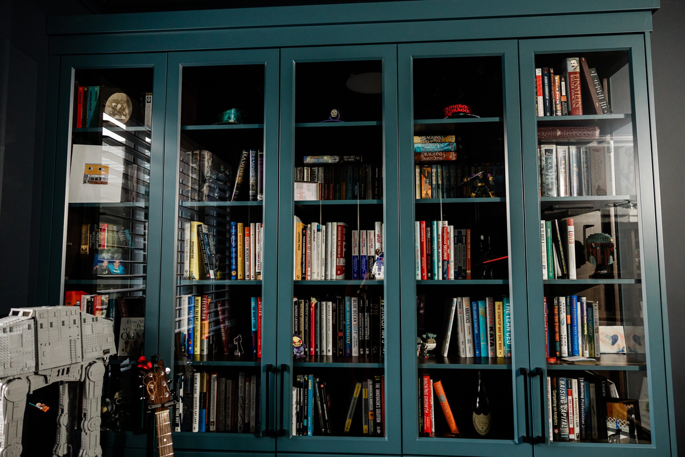 A blue bookshelf filled with books, models, and a guitar. There's a Star Wars AT-AT model on the left, suggesting a fan's collection.