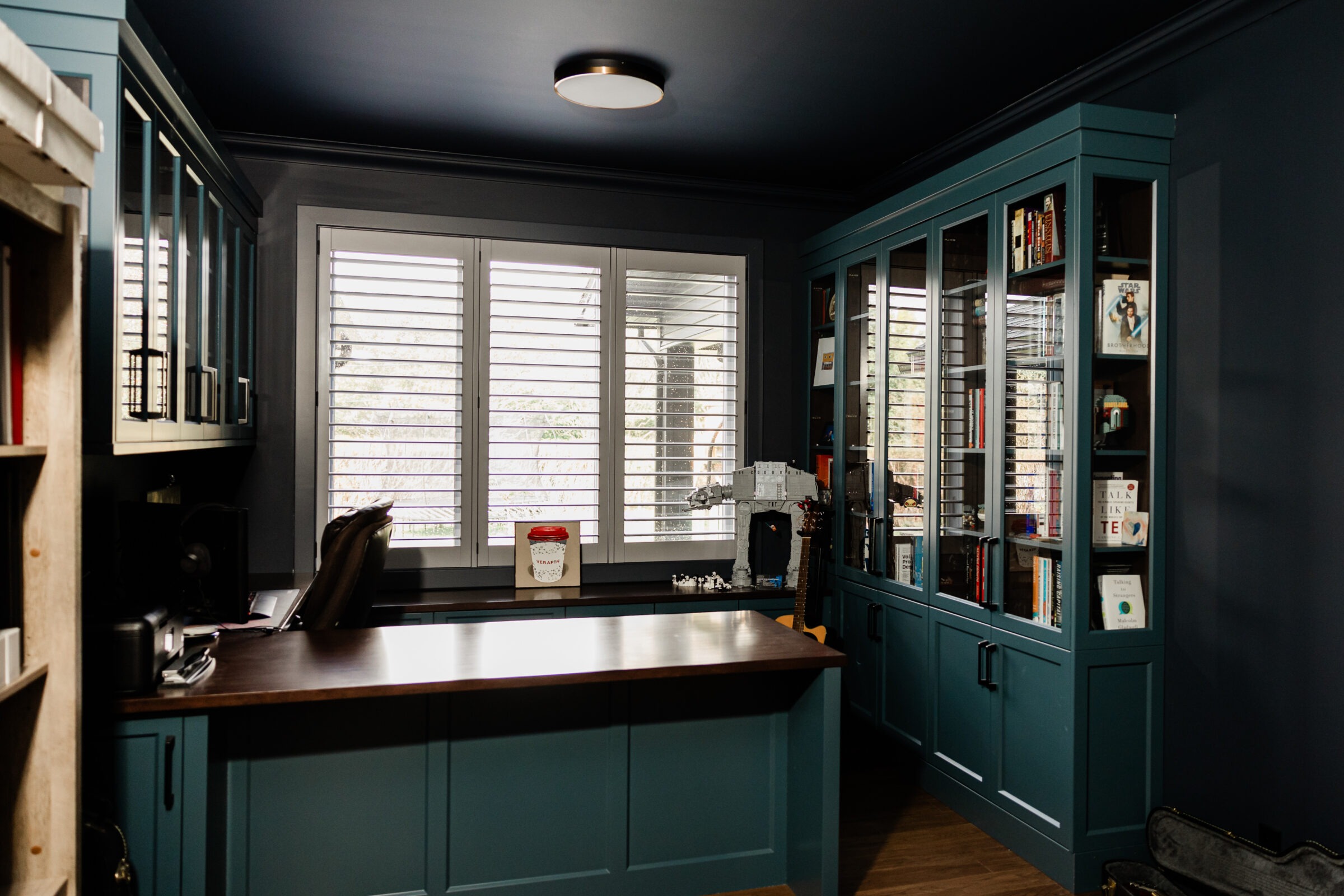 A home office with dark blue walls, teal cabinetry, and white window shutters. A desk with a computer and bookshelves filled with books and items.