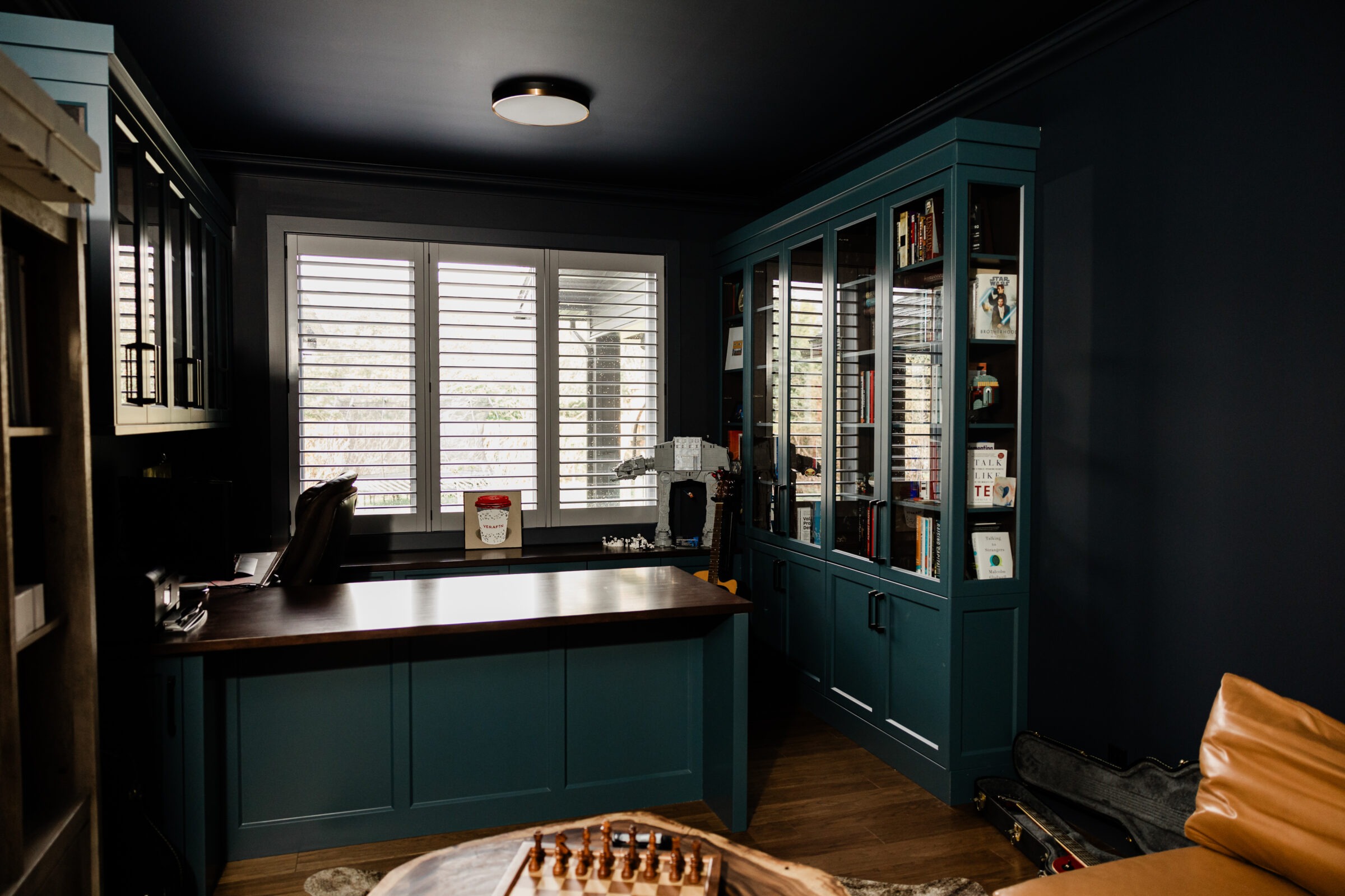 A cozy home office with dark blue walls, teal cabinetry, white shutters on windows, a desk with a computer, and a chessboard on a coffee table.