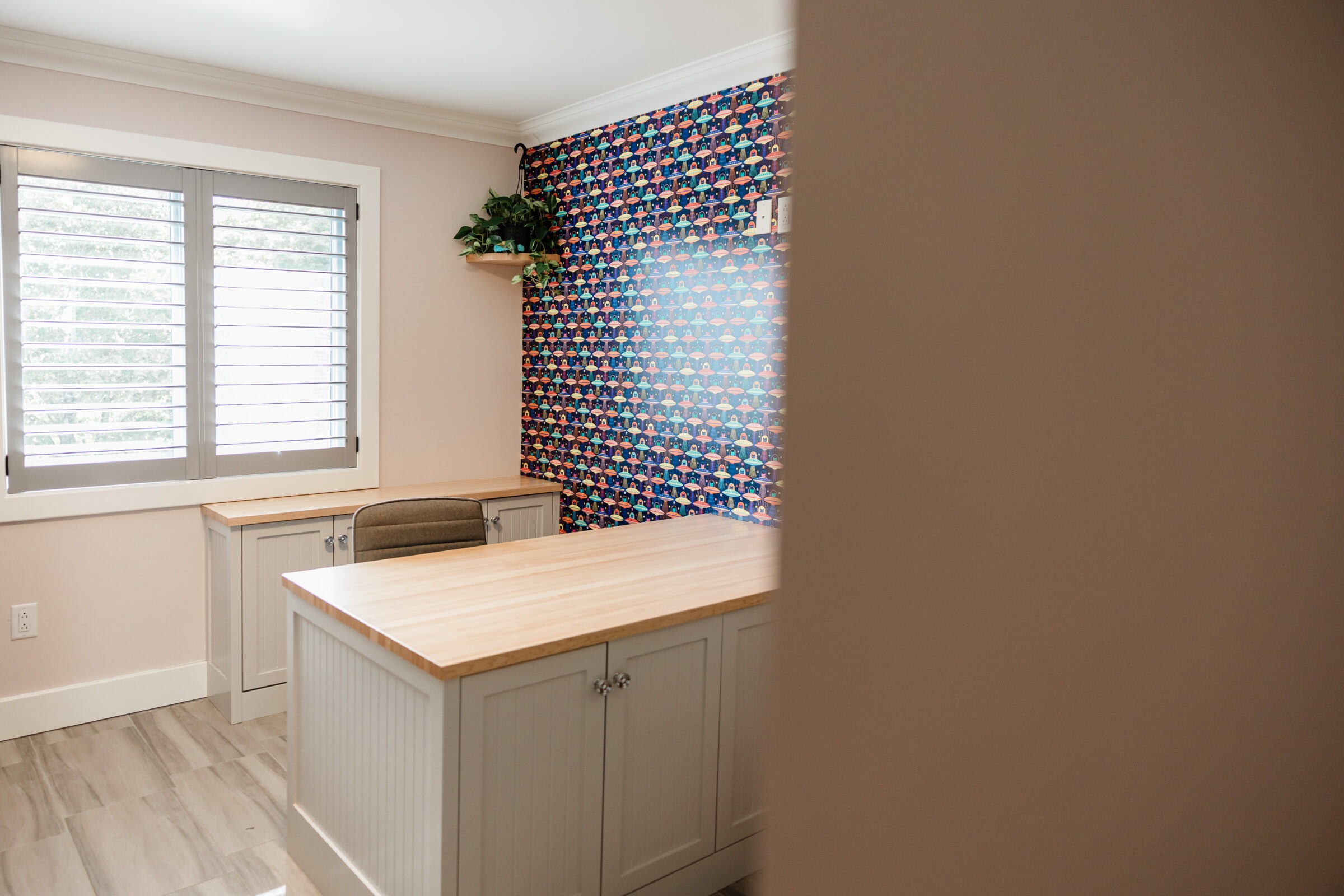 A modern kitchen corner featuring plantation shutters, light wooden countertops, white cabinetry, and a colorful tiled accent wall with a plant shelf.