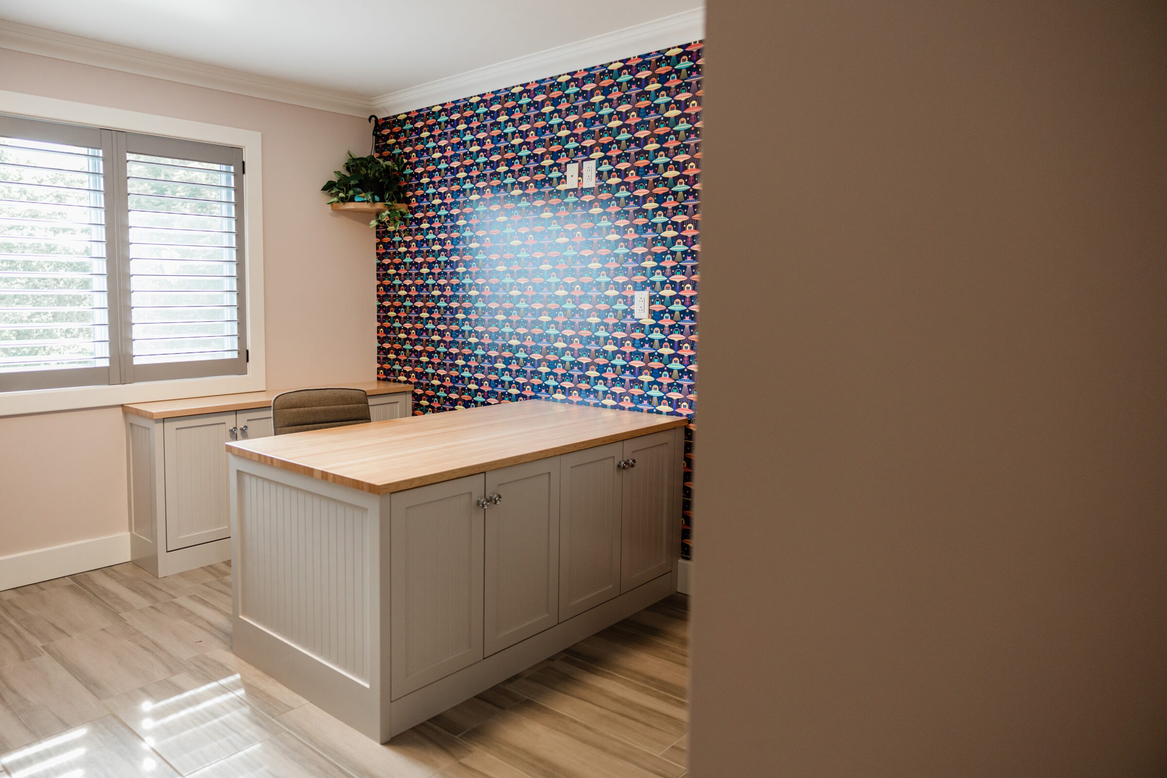 A modern room with wood flooring, a beige island cabinet, colorful patterned wall, window shutters, and a plant on a high shelf.