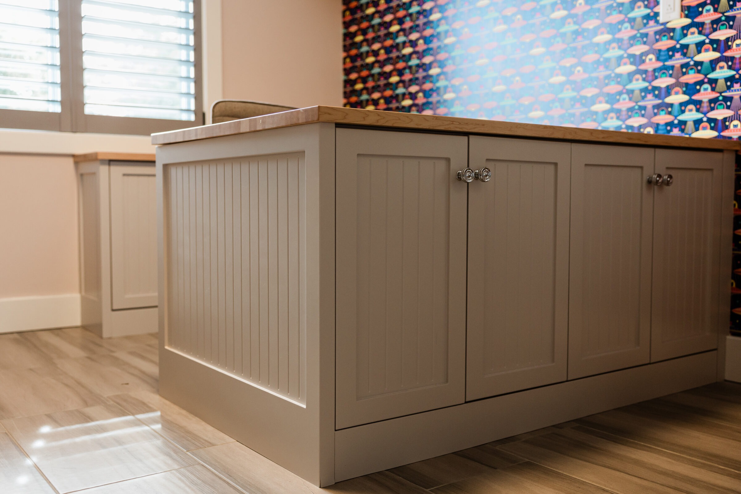 A beige cabinetry system with silver handles sits on a wooden floor in a room with a colorful geometric wallpaper and sunlight filtering through window blinds.