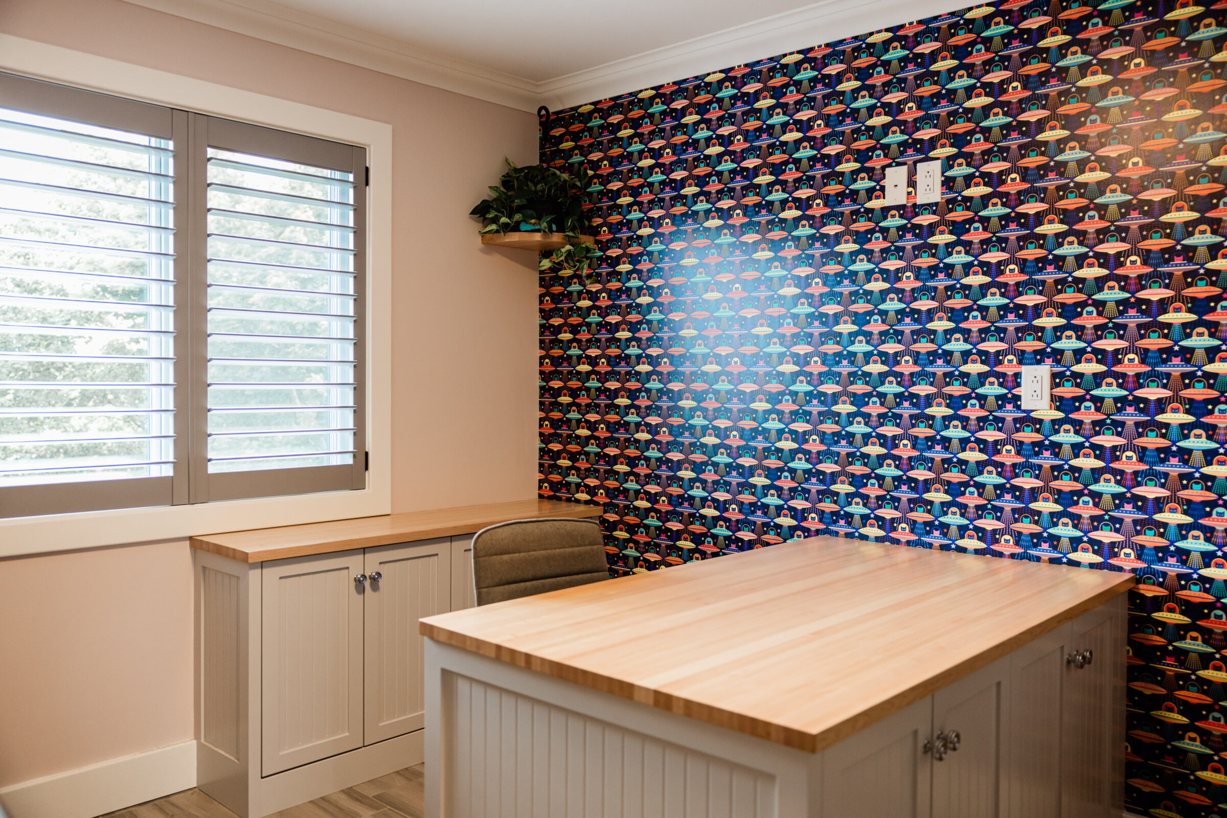 A modern office space with one wall featuring colorful, whimsical wallpaper with repeating patterns of animated characters. The room includes a desk and a chair.