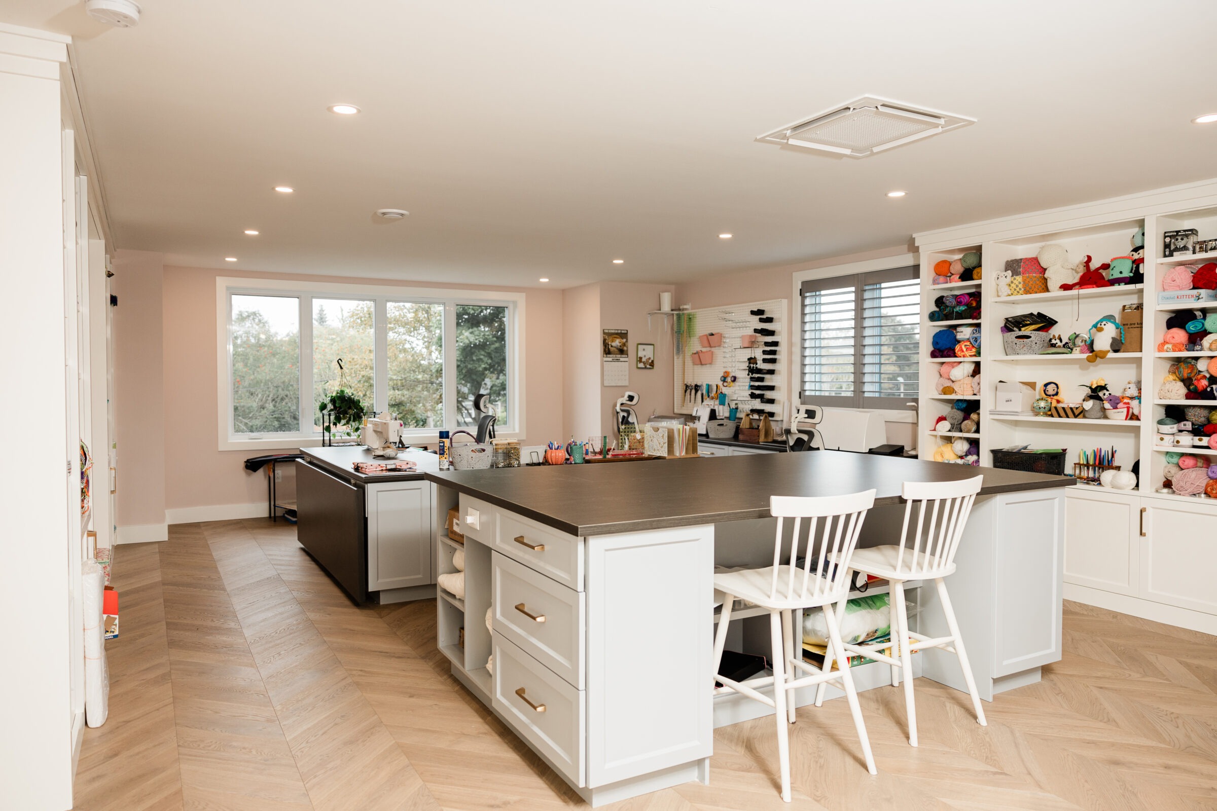 A spacious, well-lit craft room with a large central island, white chairs, ample shelf storage filled with yarn, and sewing materials on surfaces.