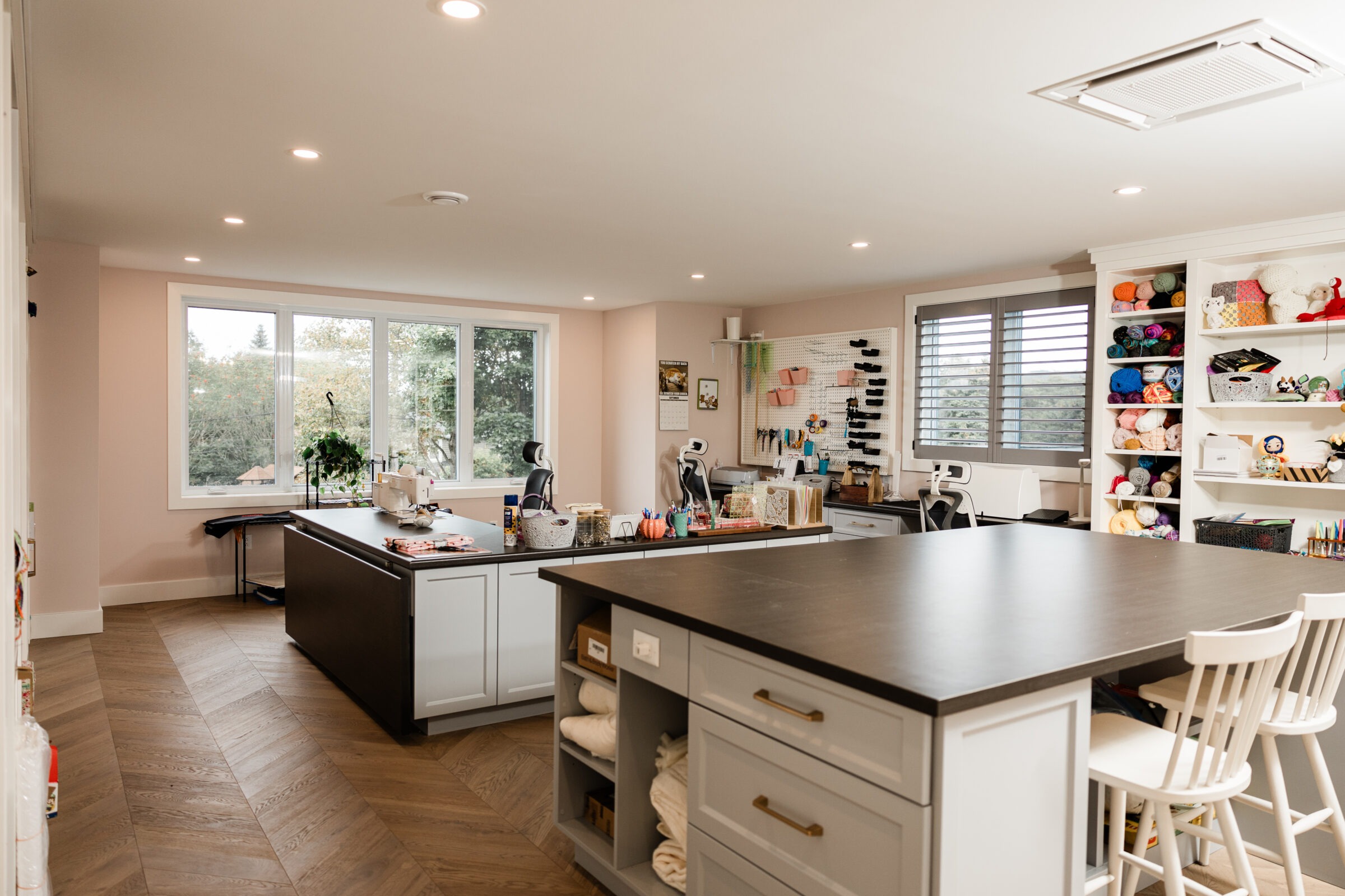 A spacious craft room with a large central island, cabinets, and storage filled with colorful yarn and crafting materials. Light fills the room from a window.