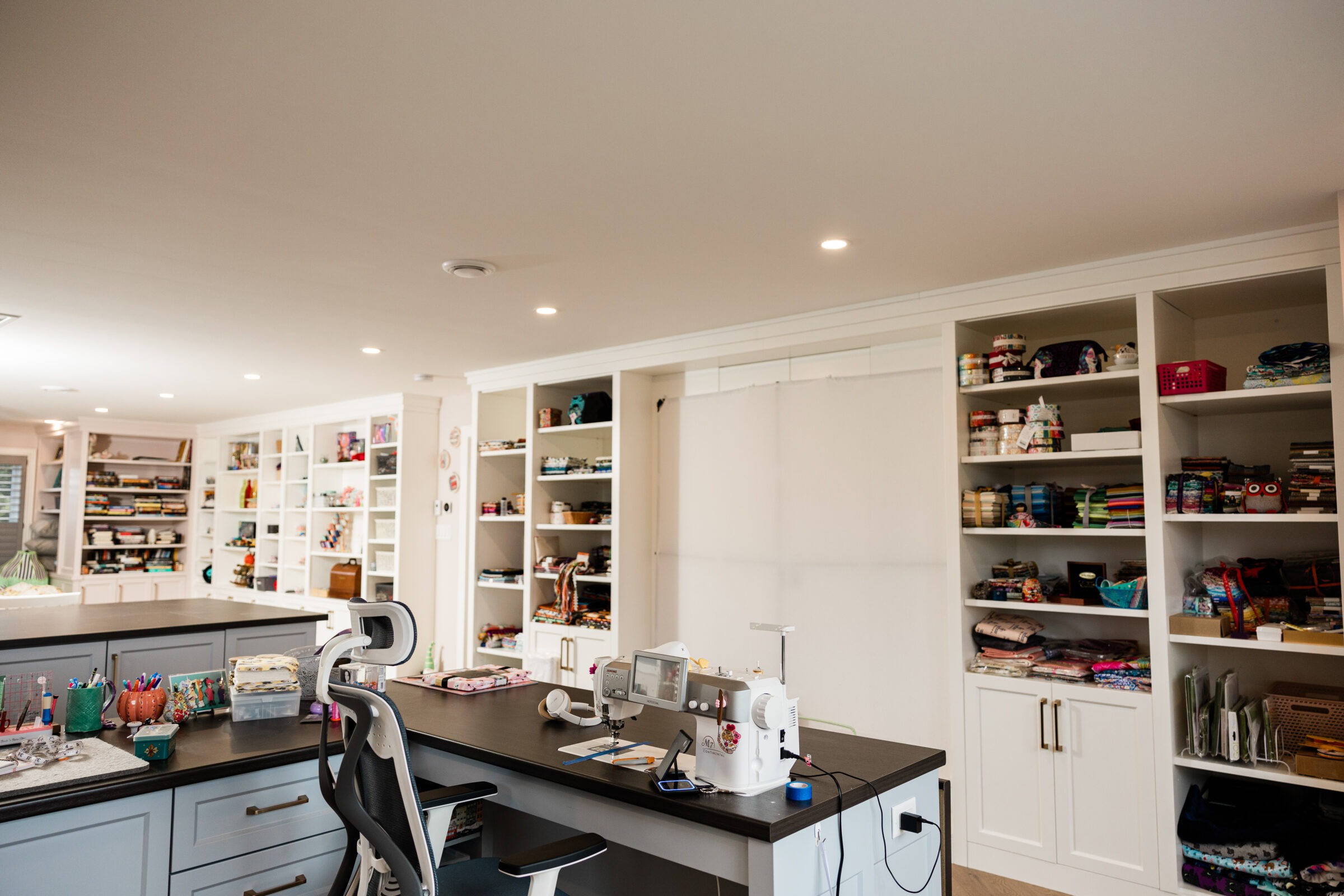 A spacious, well-lit craft room with white shelving full of supplies, a sewing station with machines, and a central island workspace.