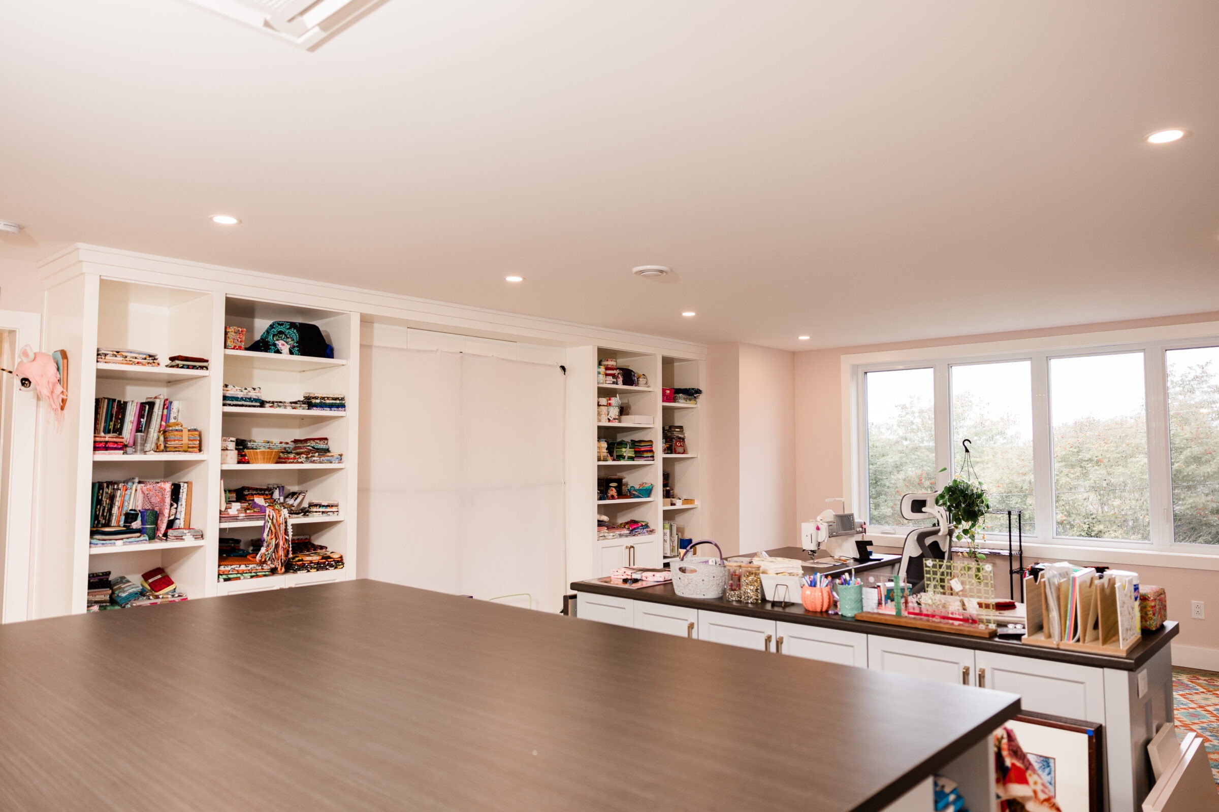 A spacious, well-lit craft room with organized shelves, a large worktable, sewing machines, and materials with a view of trees through windows.