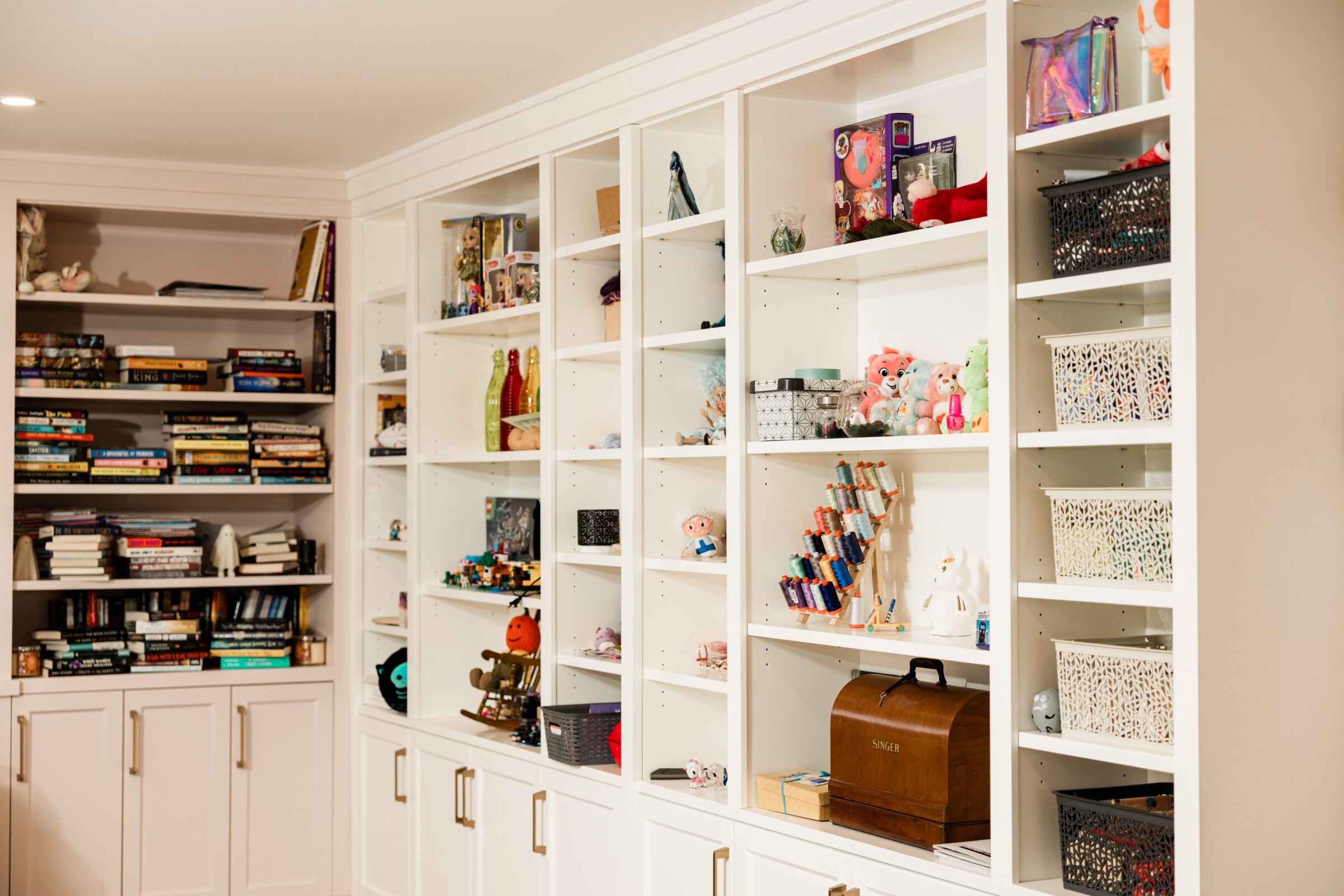 A spacious, white built-in shelving unit containing books, toys, sewing threads, and decorative items, with a vintage Singer sewing machine prominently displayed.
