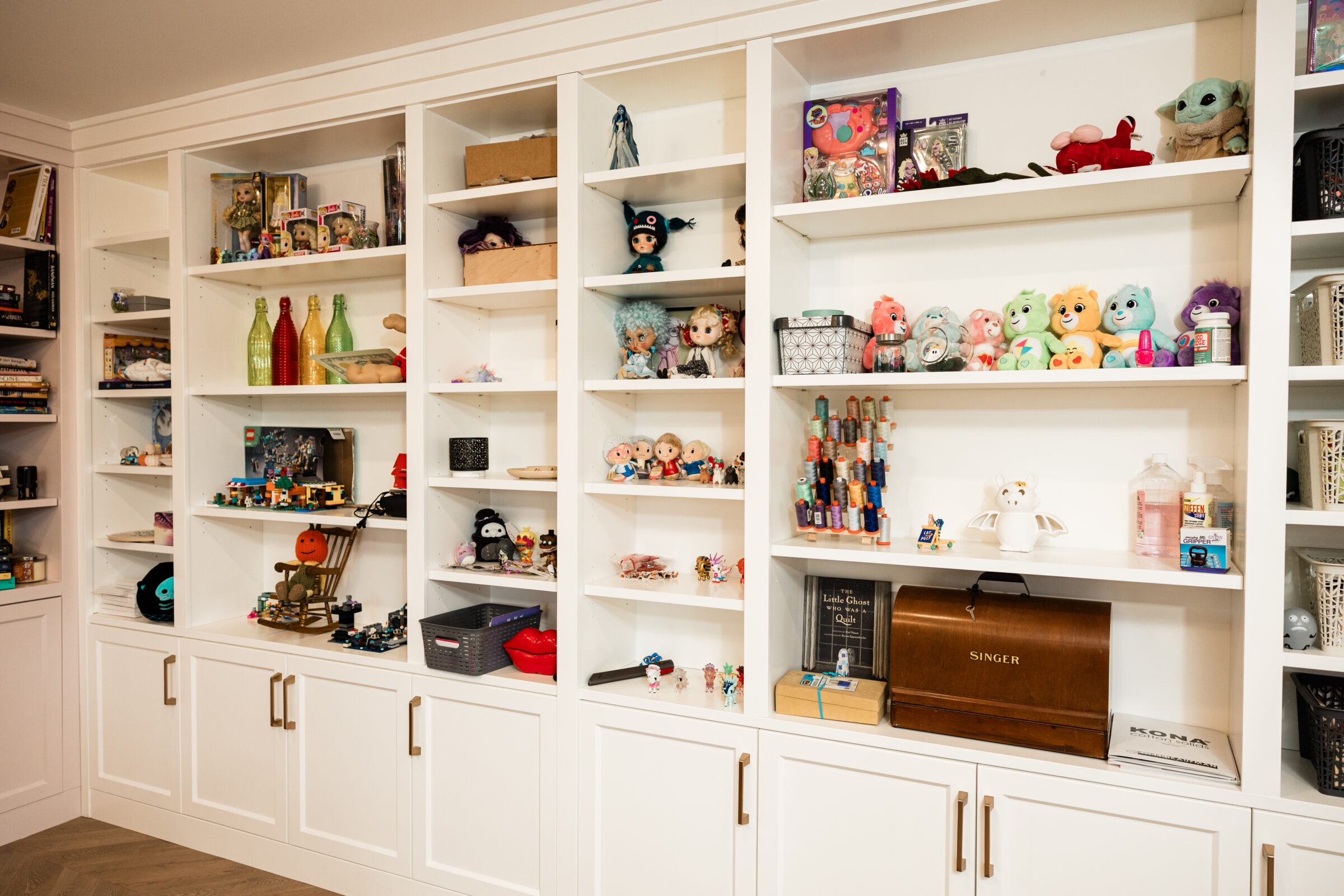 White built-in shelves are filled with toys, dolls, bottles, books, and a vintage sewing machine, reflecting a cozy, organized, hobby or craft room.