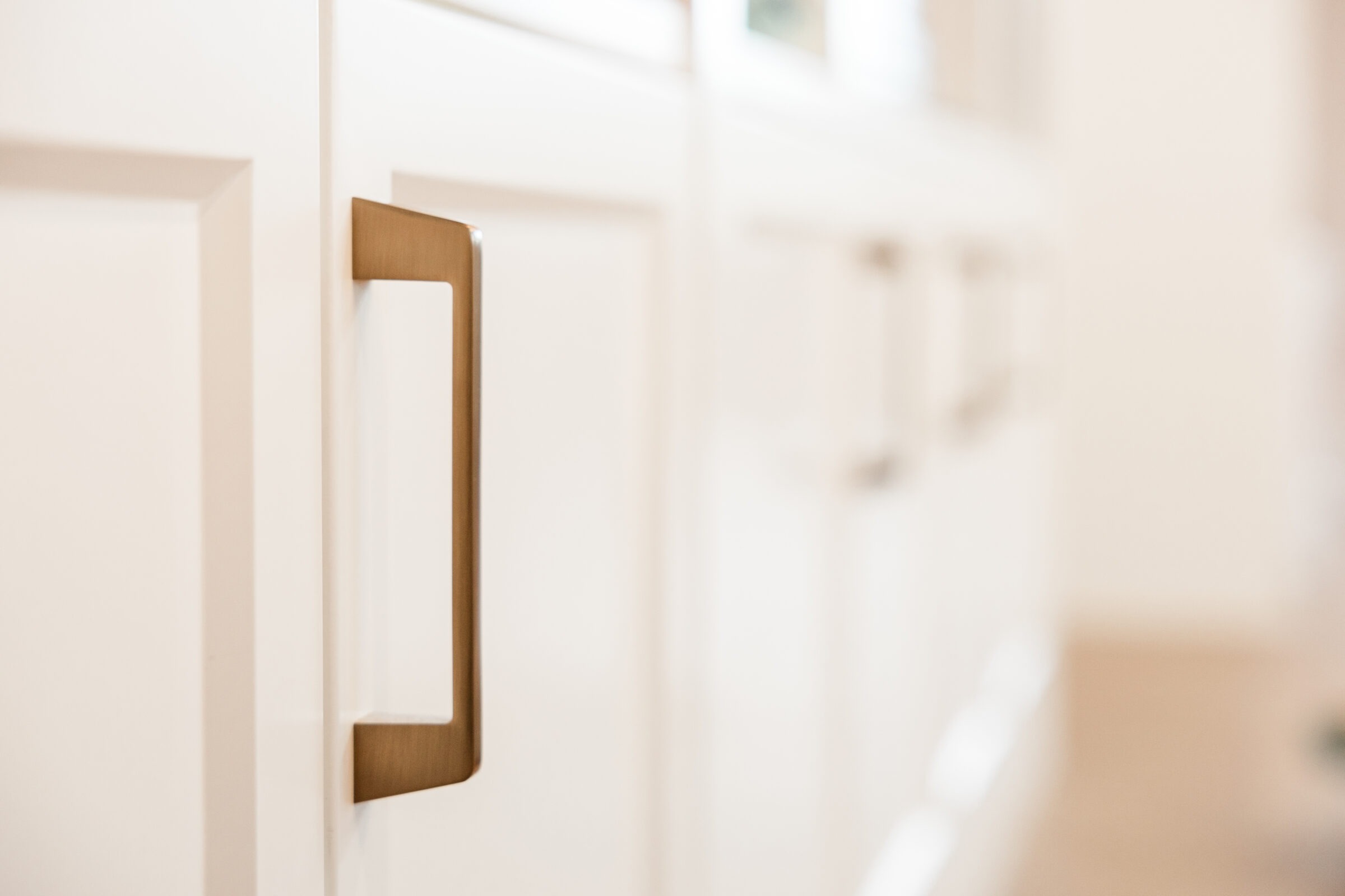 A close-up of a modern bronze cabinet handle on a white shaker-style cabinet door, with blurred cabinetry in the background, suggesting a clean, minimalist kitchen.