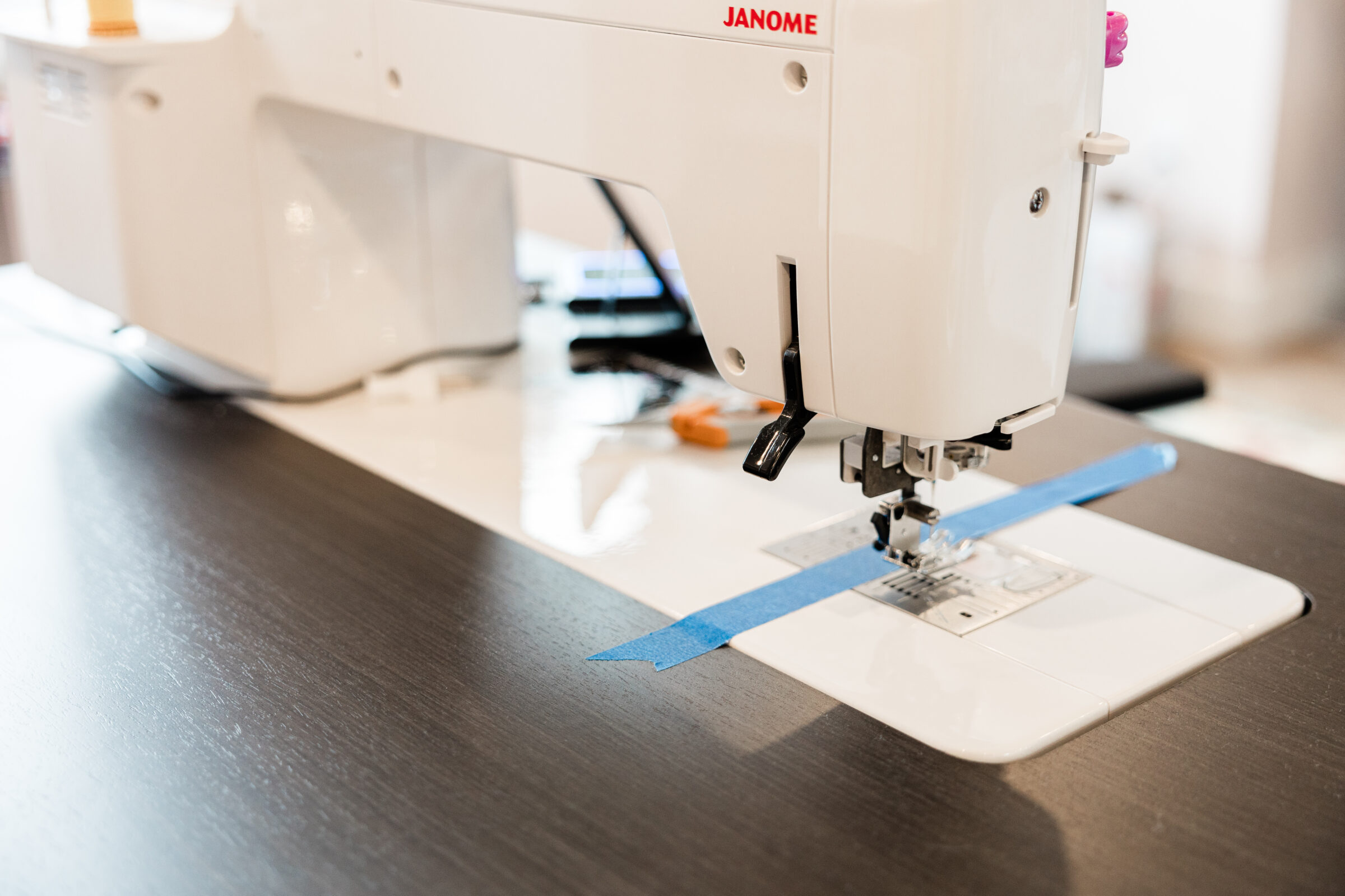 This image shows a close-up of a white Janome sewing machine on a dark surface, featuring a needle area and blue guiding tape.