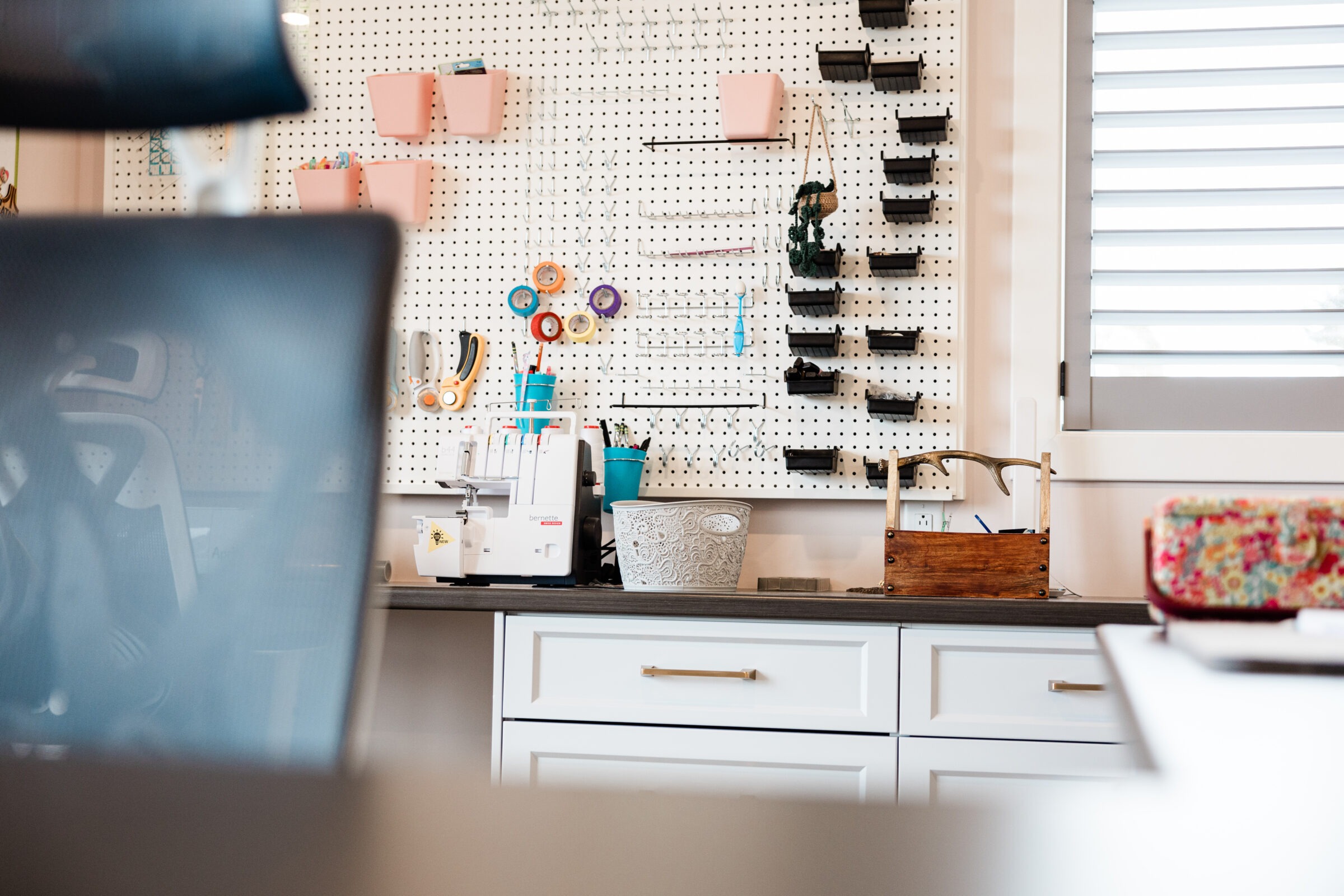 A well-organized craft room with a pegboard tool organizer, white cabinetry, a sewing machine, and scissors. A blurred foreground suggests a work session.