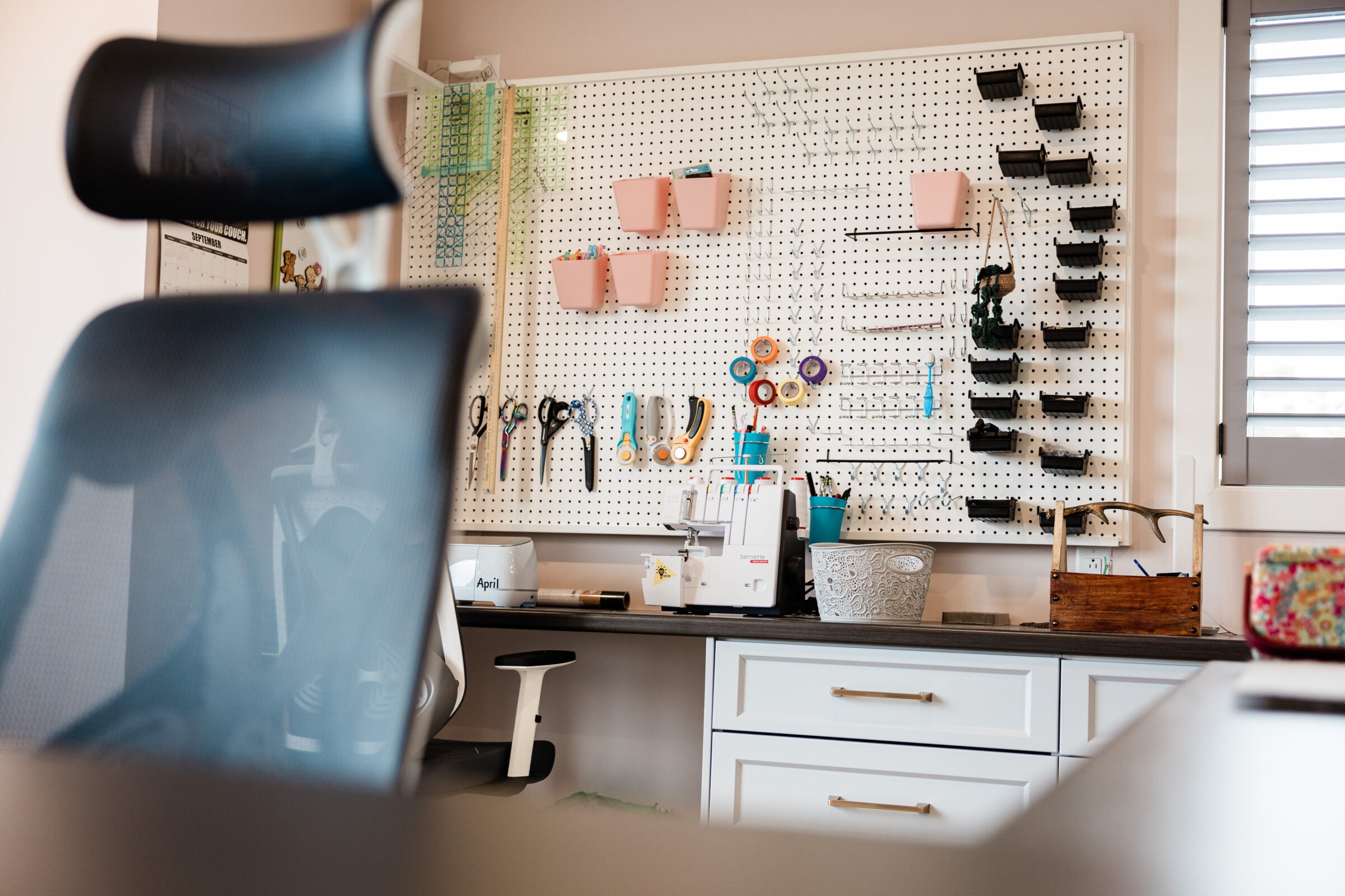 An organized craft room with a pegboard holding tools, scissors, and tape. A sewing machine, calendar, and desk chair are visible. Warm, inviting atmosphere.
