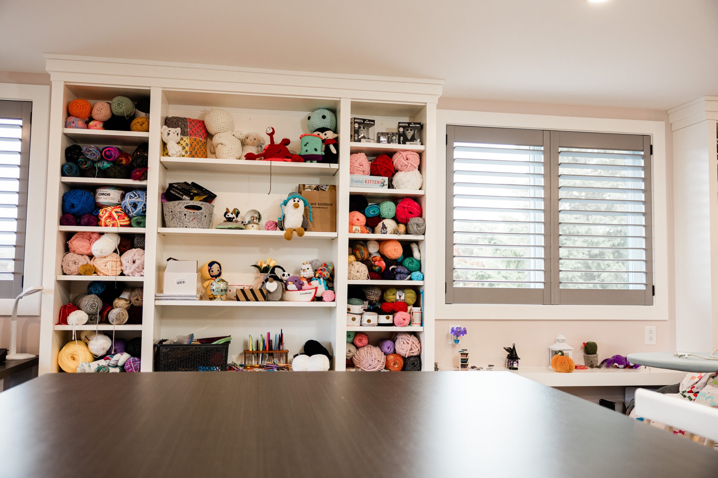 A craft room with neat shelves filled with colorful yarn balls, handcrafted toys, and crafting materials. A large table is in the foreground.