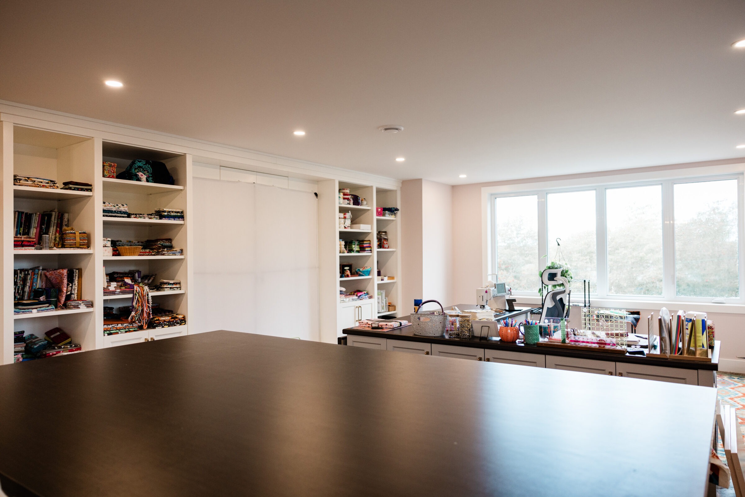 A spacious room with ample natural light, featuring a large black tabletop, shelving with books and fabric, and a sewing station by the window.