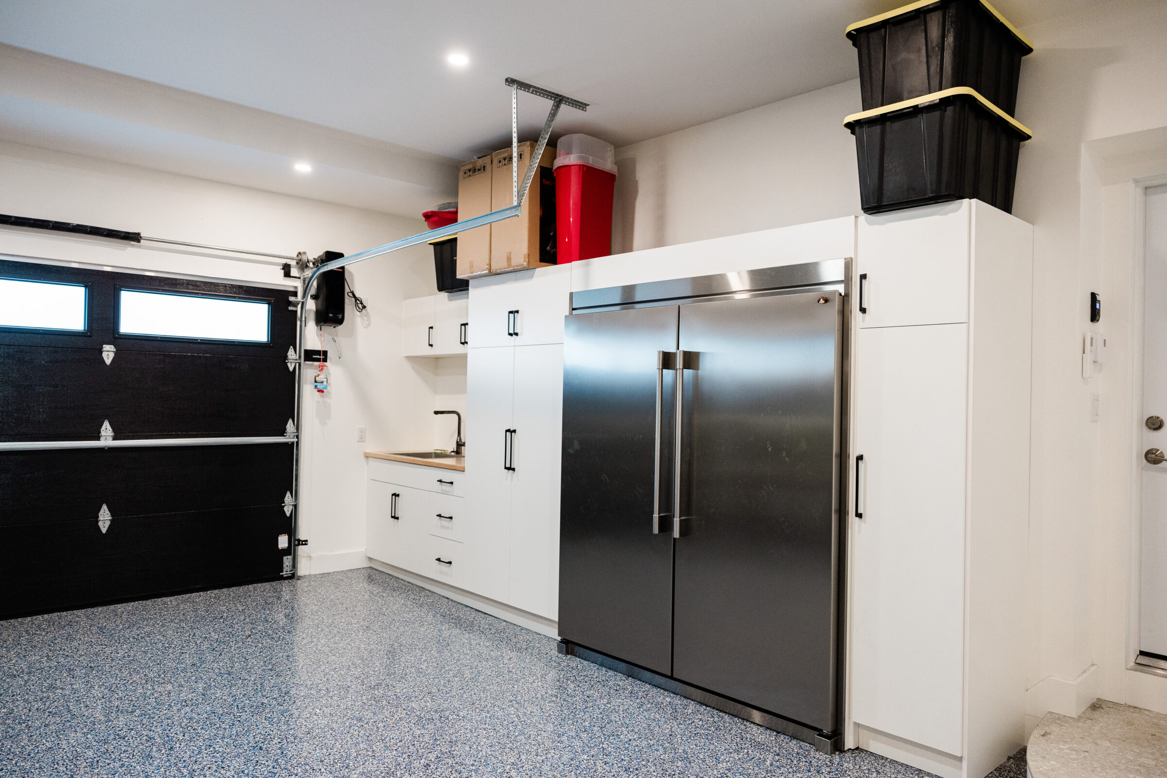A clean, modern garage interior with white cabinetry, stainless steel fridge, speckled floor, black garage door, and overhead storage solutions.