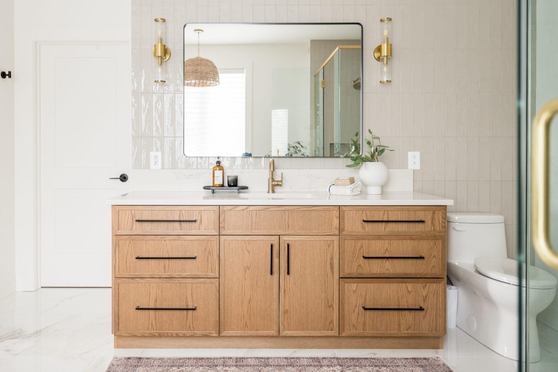 Modern bathroom interior with wooden vanity cabinet, large mirror, brass fixtures, white countertop, decorative plants, and a visible toilet area.