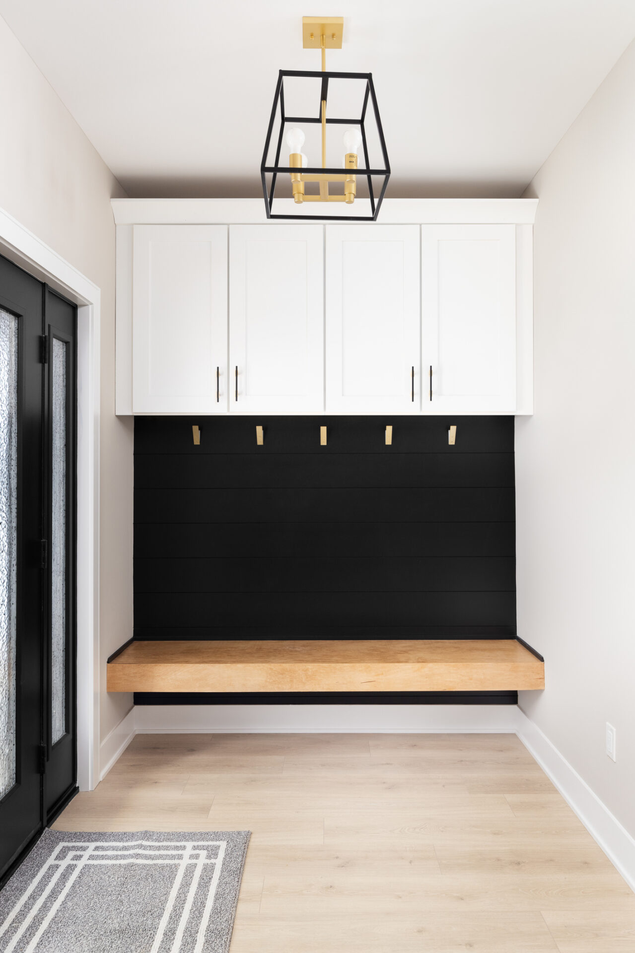 Modern interior entryway featuring white upper cabinetry, black shiplap wall, floating wooden bench, geometric chandelier, light wood floor, and a gray mat.