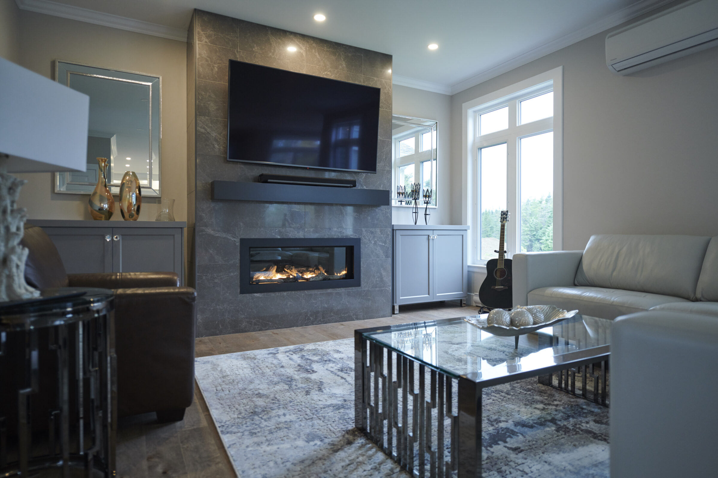 A modern living room featuring a fireplace, mounted TV, white sofa, glass coffee table, decorative vases, and a guitar near bright windows.