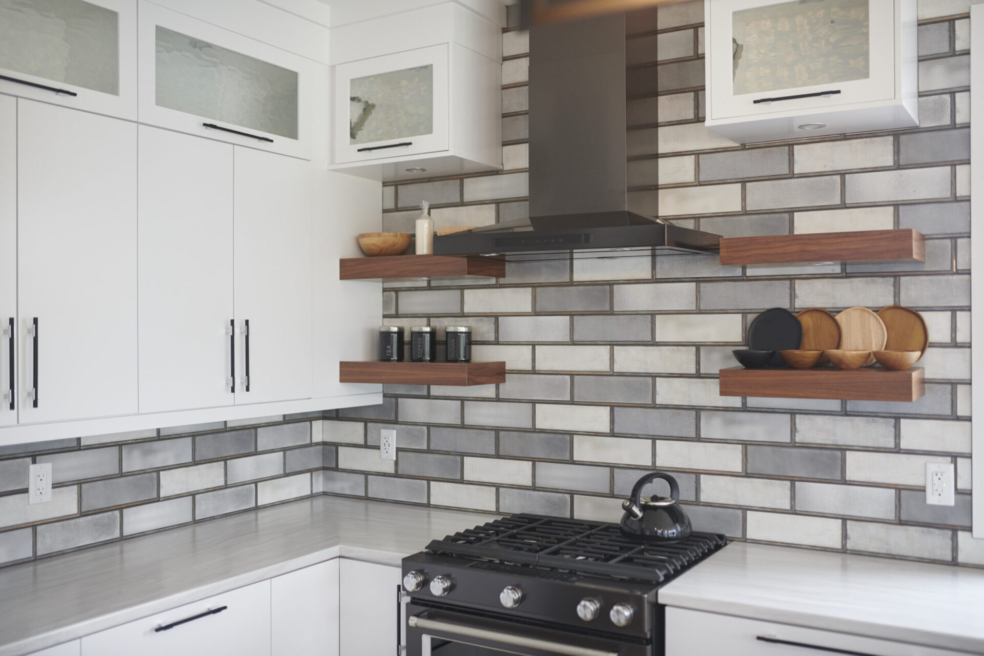 A modern kitchen with white cabinets, stainless steel appliances, gray backsplash tiles, wooden shelves, and minimalist decor. There's a black gas stove and kettle.