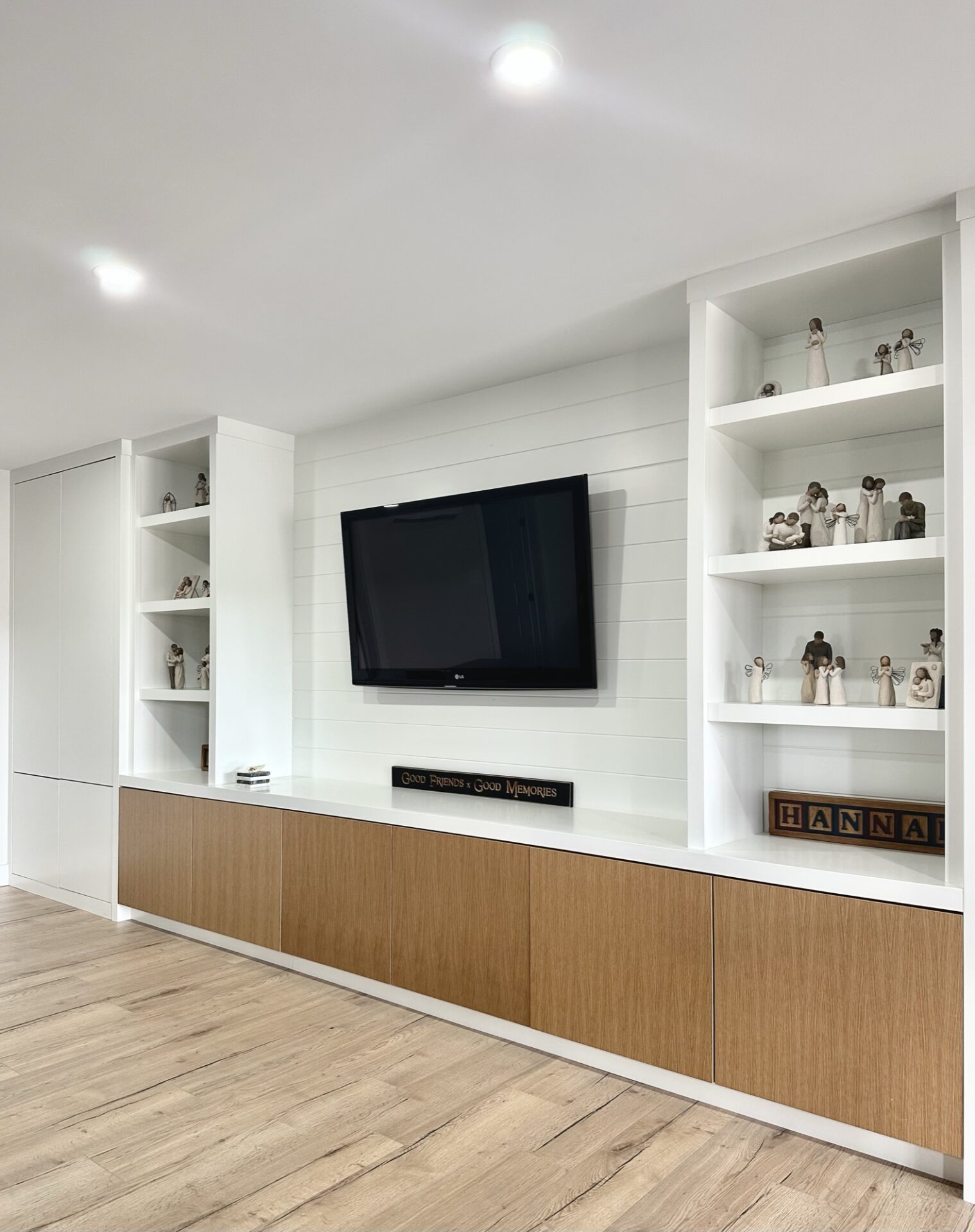 Modern living room interior featuring a mounted TV between white shelving units displaying decorative figures, with a wooden cabinet below and recessed lighting above.