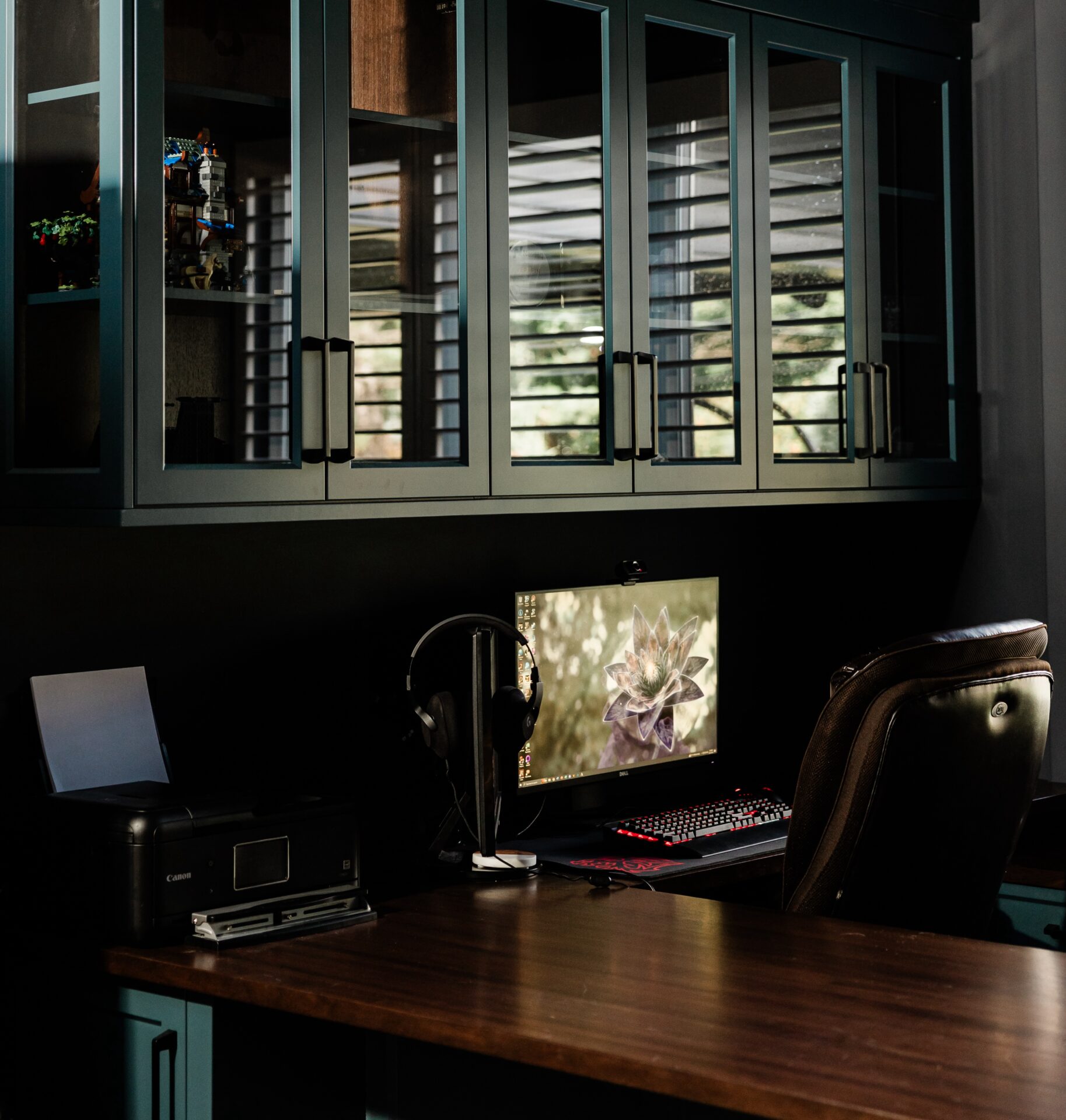 This is a home office setting featuring a wooden desk with a laptop, printer, computer with dual monitors, and headphones. Sunlight filters through window blinds.