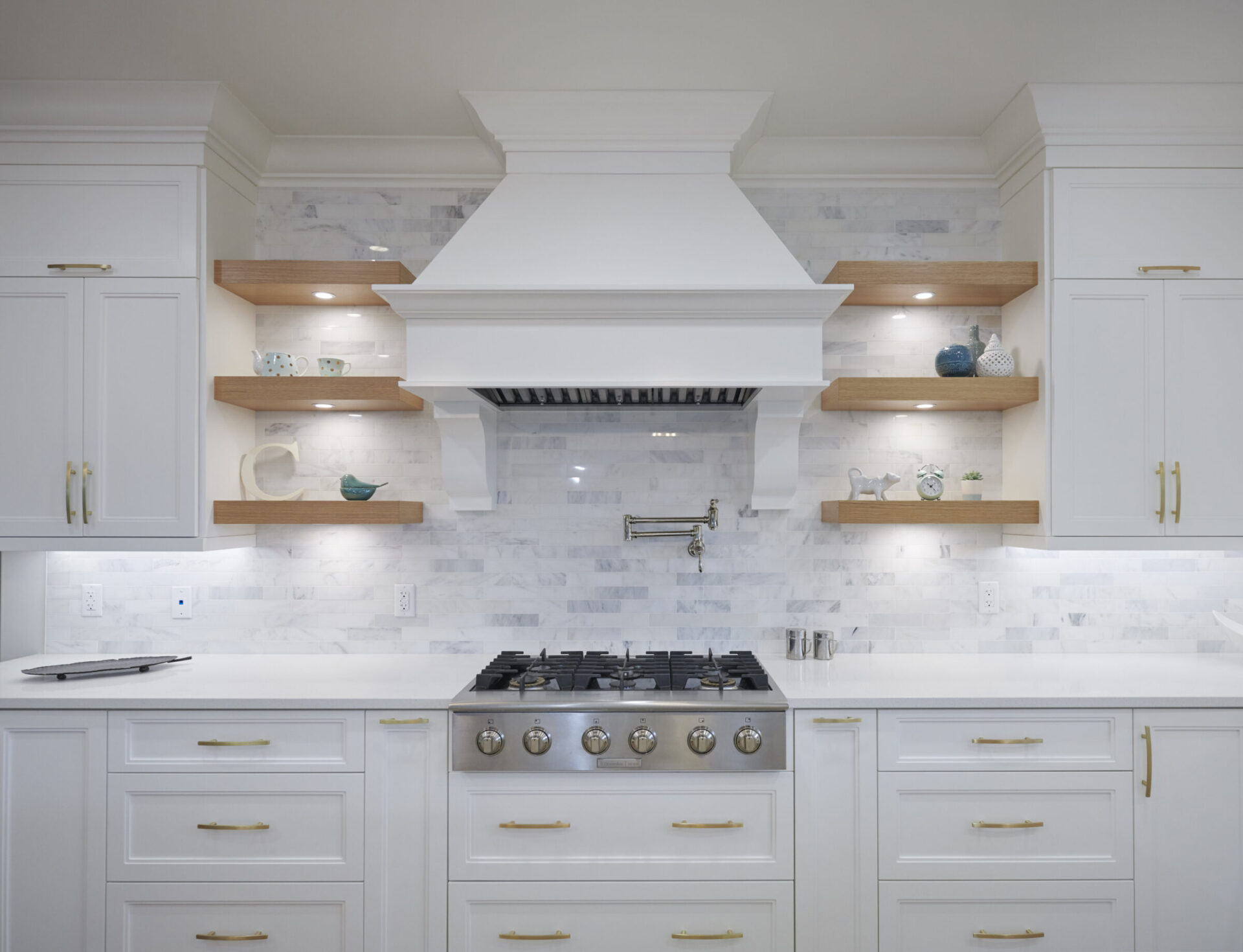 A modern kitchen with white cabinetry, marble backsplash, wooden open shelves, brass hardware, and a stainless steel range with a range hood.