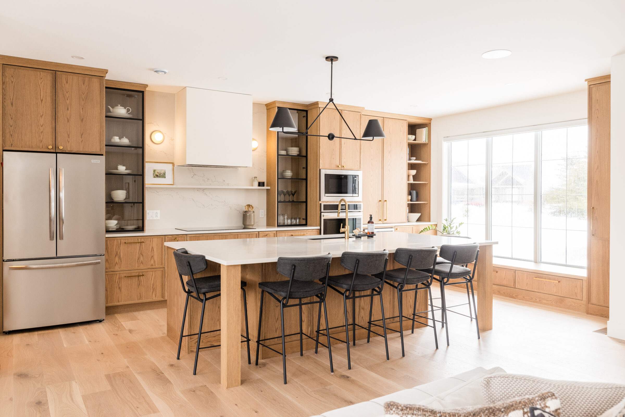 A modern, well-lit kitchen with wood cabinets, a central island with bar stools, stainless steel appliances, and a large window with a snowy view.