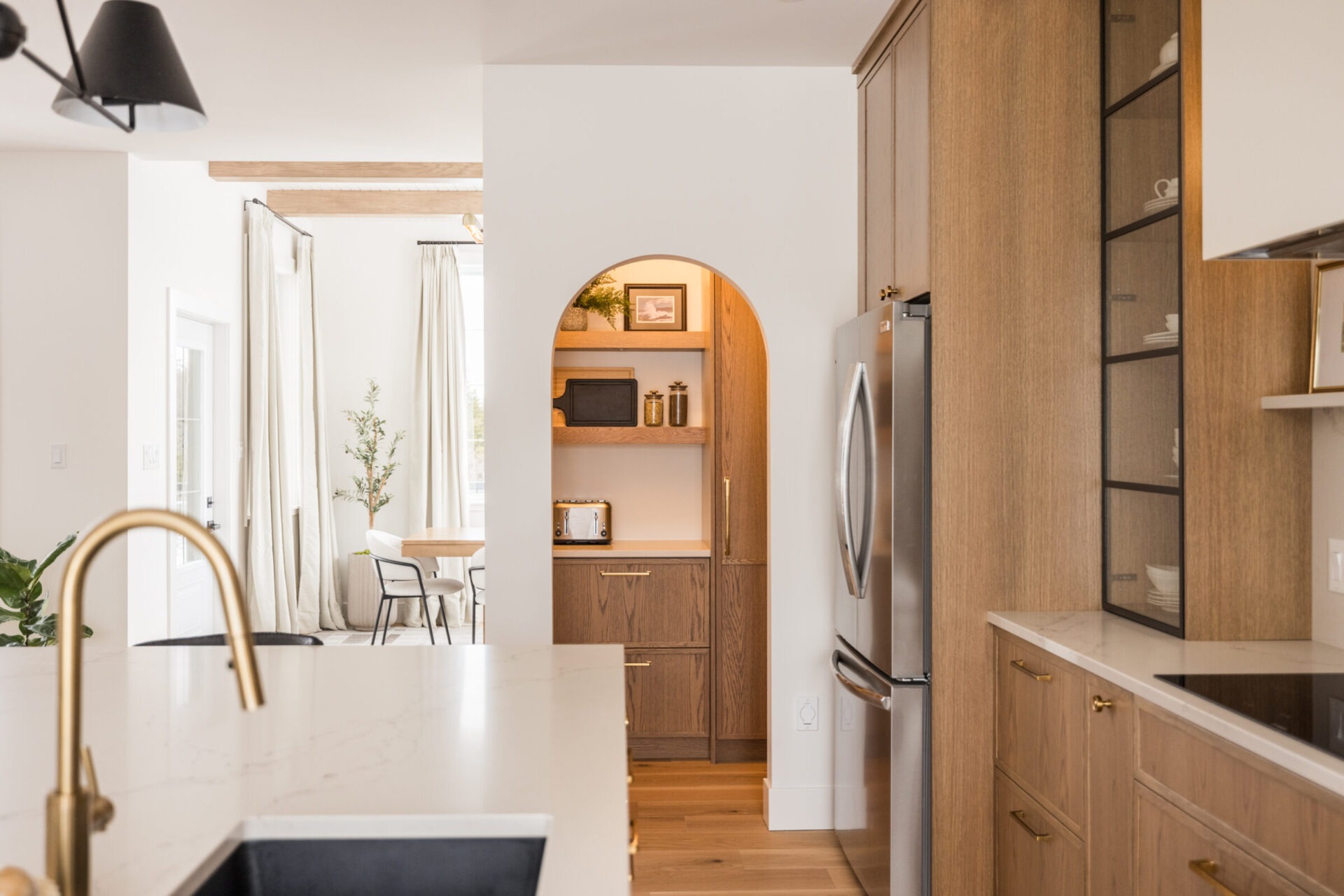 The image displays a modern kitchen with wooden cabinetry, a marble countertop, a gold faucet, stainless steel appliances, and an arched doorway.
