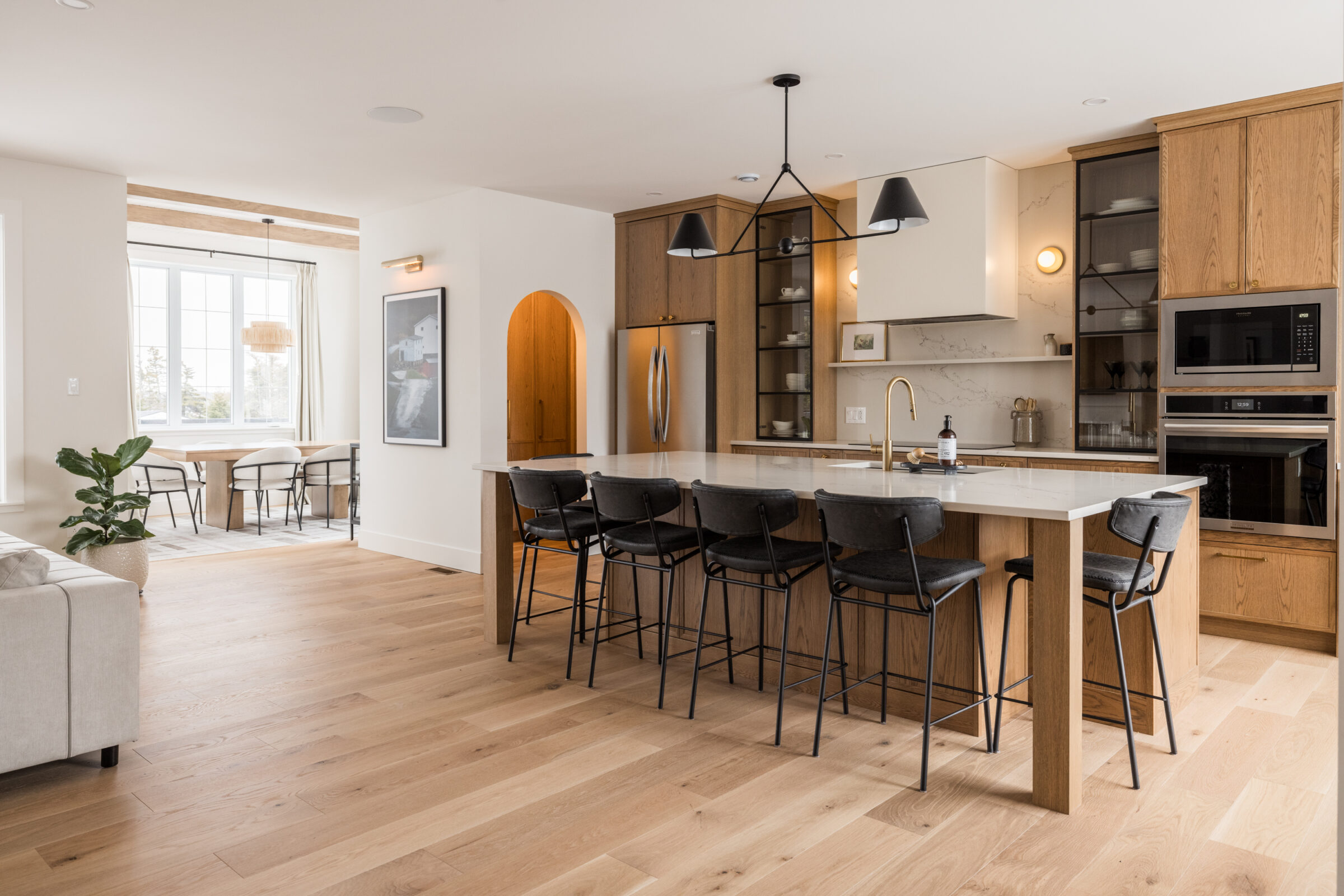 Modern kitchen interior with wooden cabinets, white countertops, bar stools, and integrated appliances. A dining area and living room are visible in the background.