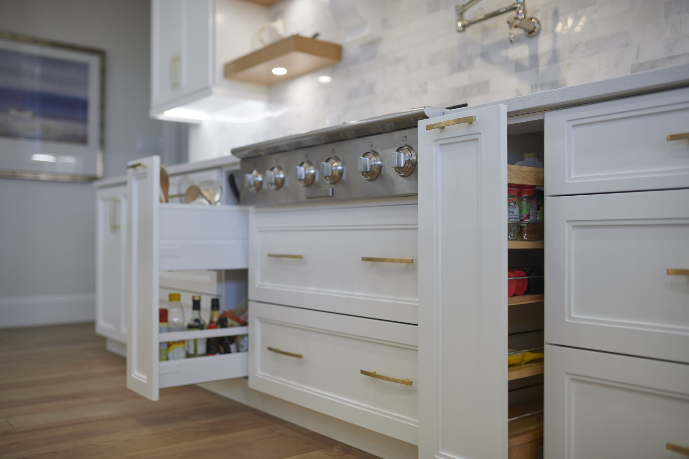 A modern kitchen with white cabinetry, gold handles, and a stainless steel stove. Open drawers reveal organized spices and utensils. Herringbone tile backsplash is visible.
