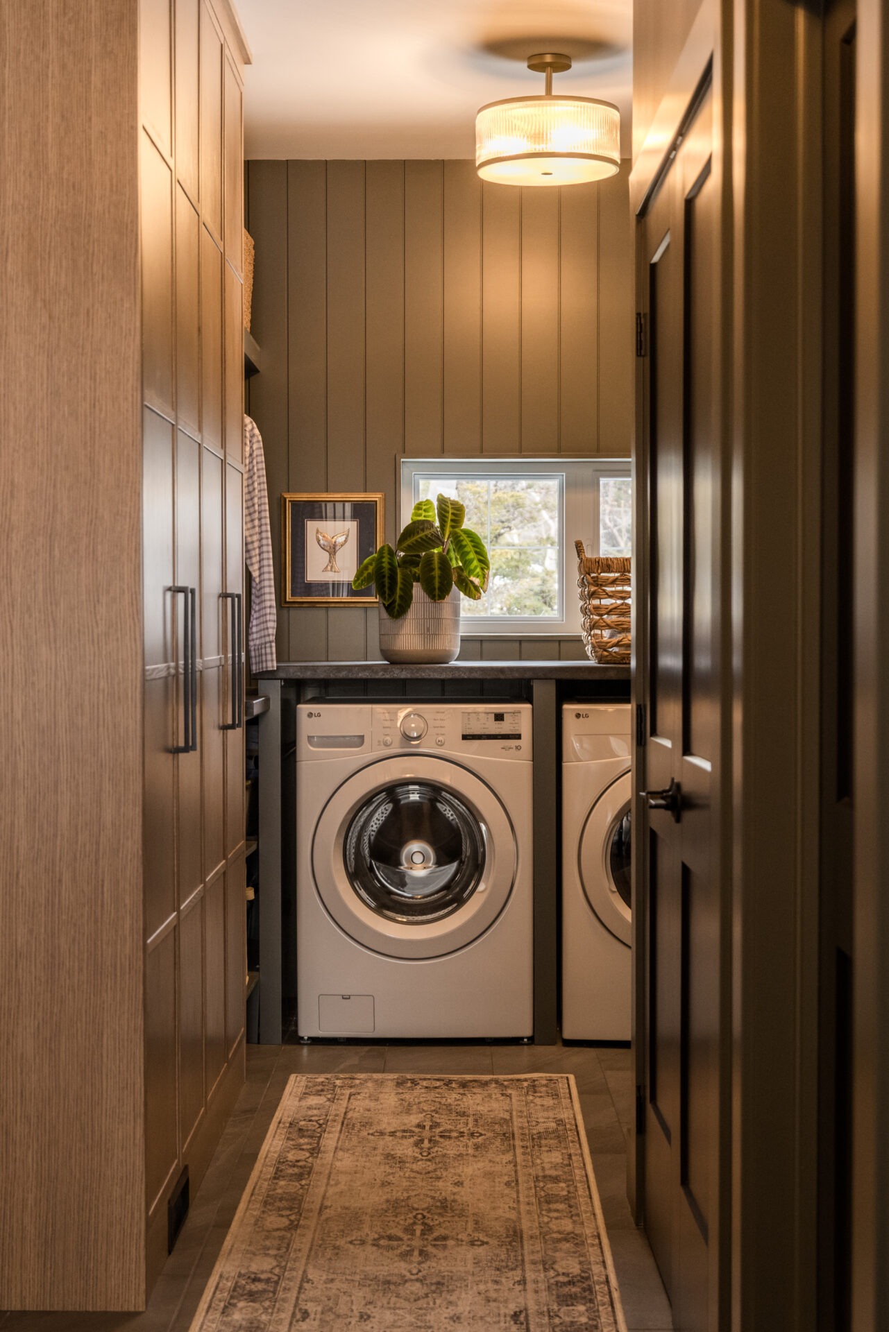A cozy laundry room with a front-loading washer and dryer, wooden cabinetry, a patterned rug, pendant light, and a window with greenery outside.
