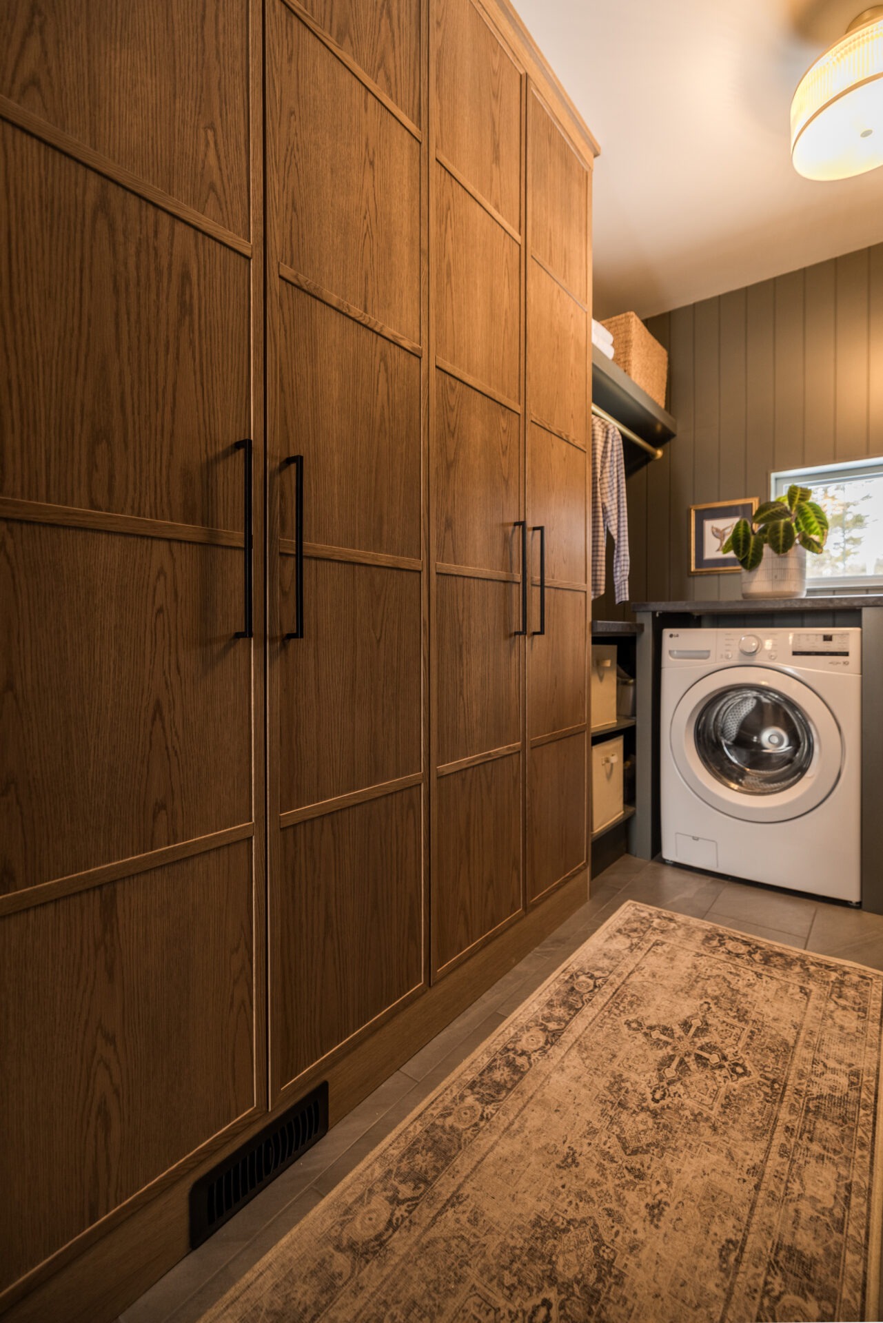 An interior space featuring tall wooden cabinetry with black handles, a front-loading washing machine to the right, a patterned rug on the floor, and a small window.