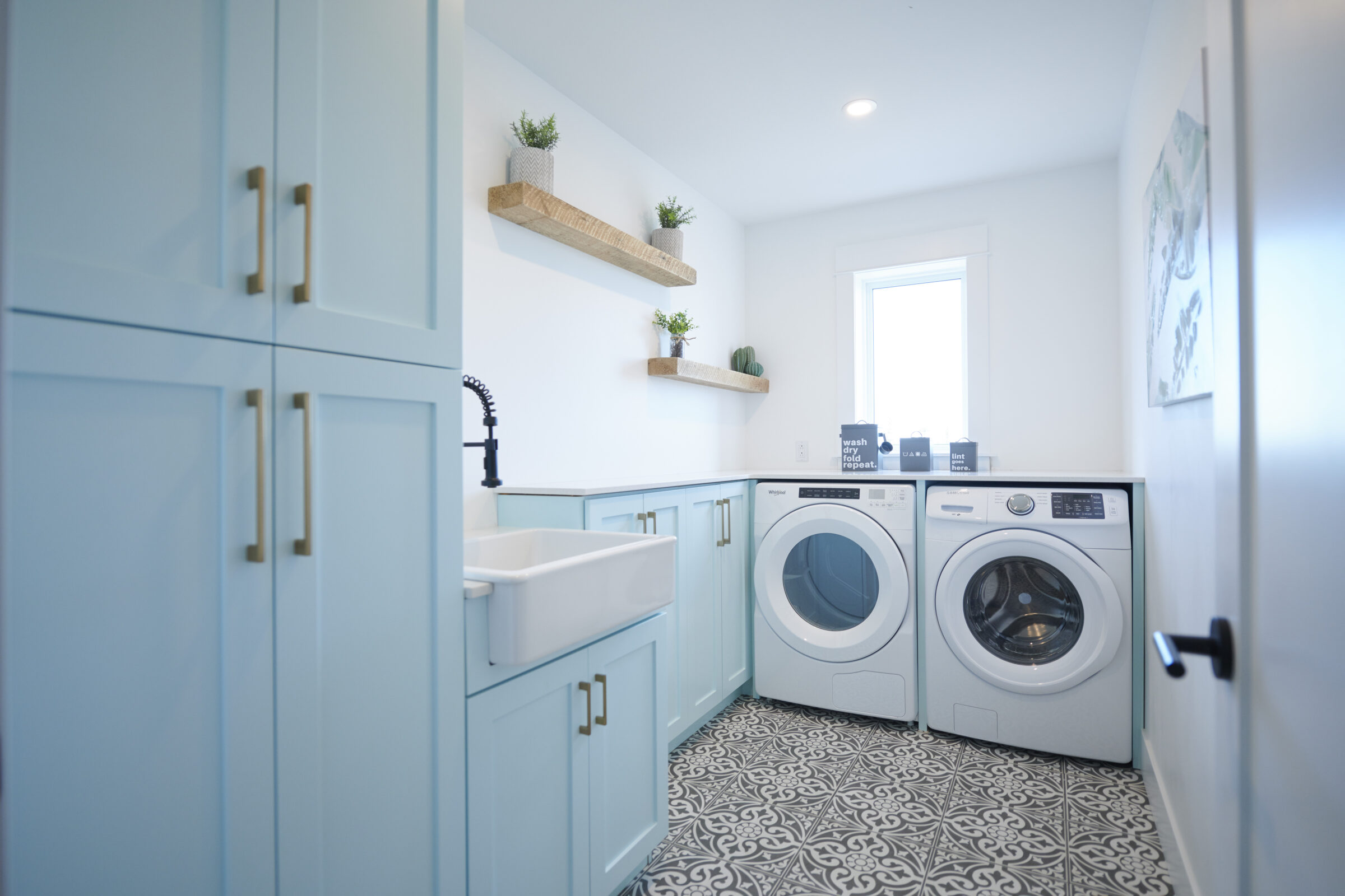 A bright laundry room with light blue cabinets, a white utility sink, washer, dryer, and floating shelves on a patterned floor.