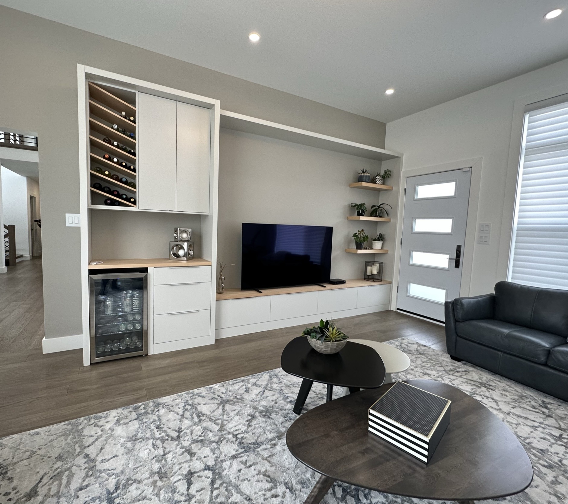 Modern living room with built-in white cabinetry, wine rack, beverage cooler, flat-screen TV, gray couch, decorative shelves, and a contemporary rug.