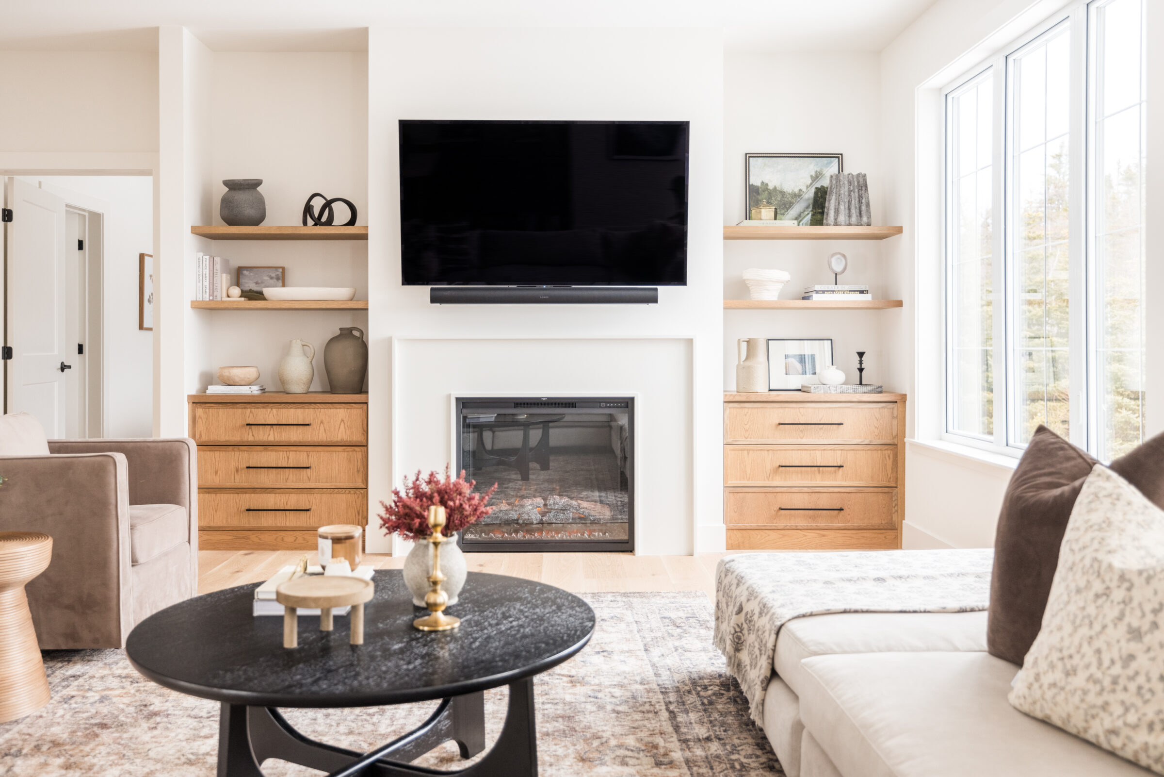 A modern living room with neutral tones, featuring a fireplace, flat-screen TV, wooden furniture, and abundant natural light from a large window.
