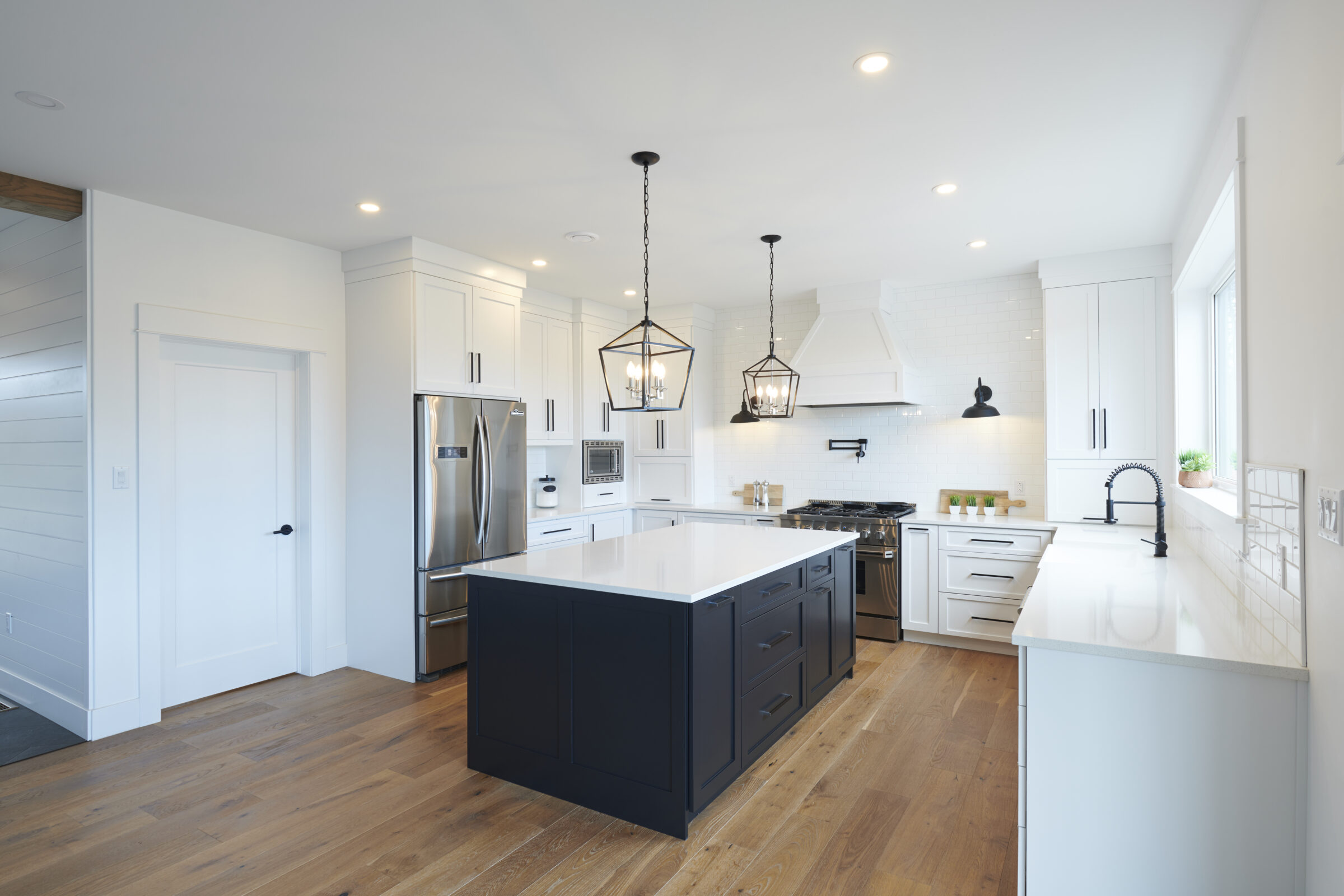 Spacious, modern kitchen with white cabinetry, dark island, stainless steel appliances, hardwood floors, pendant lights, and a subway tile backsplash.
