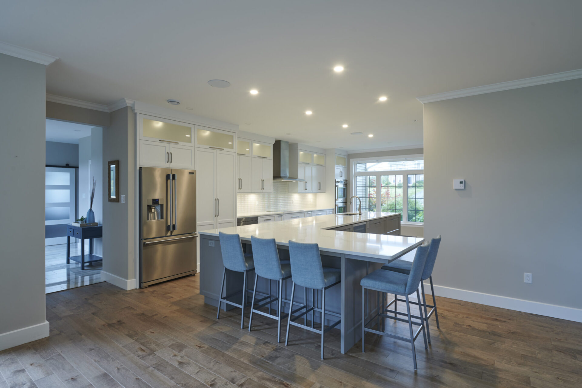 Modern kitchen interior with white cabinets, stainless steel appliances, blue chairs at a central island, hardwood floors, and recessed lighting. Large windows provide natural light.