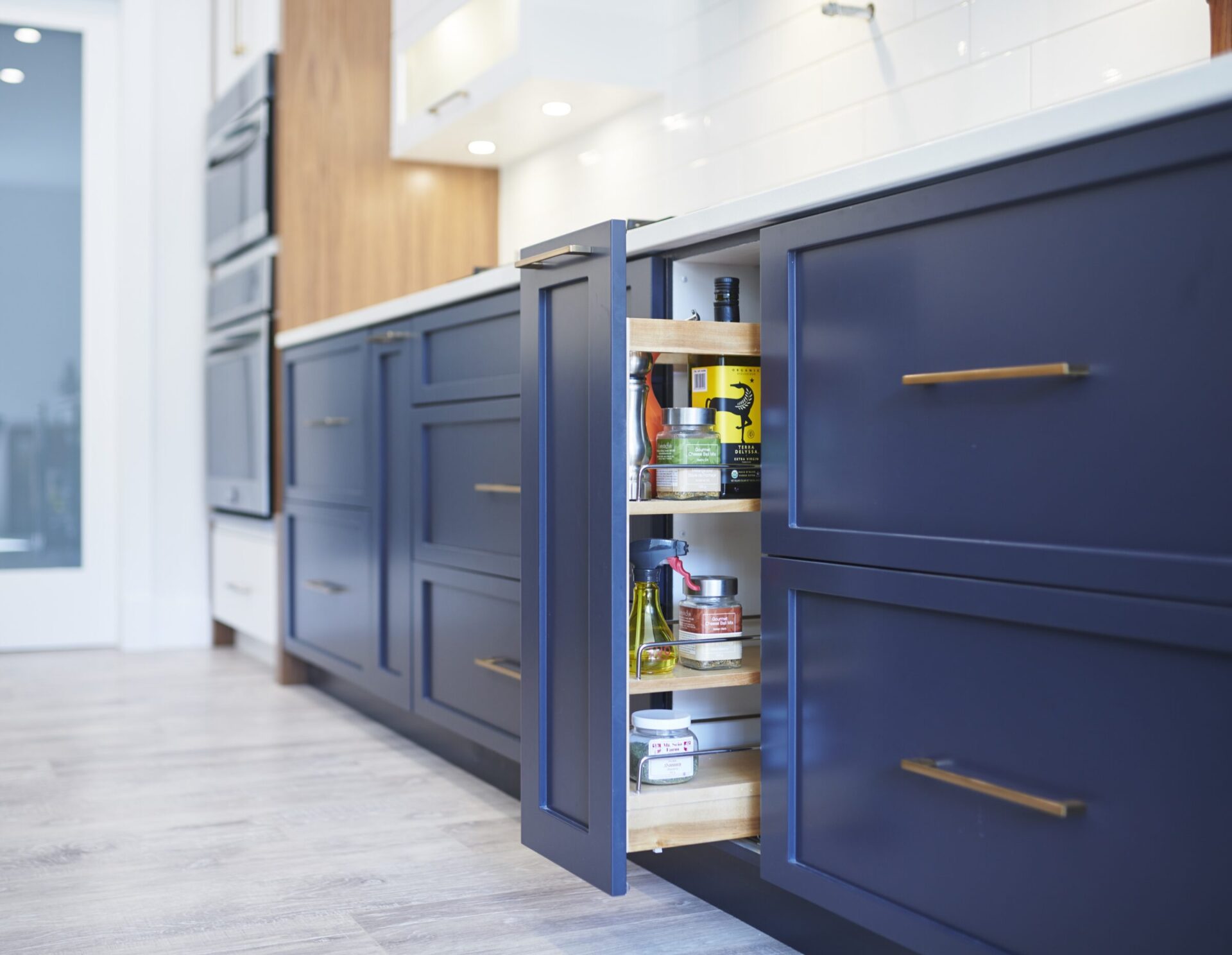 A modern kitchen interior with navy blue cabinets, a pull-out storage drawer opened to reveal bottles and jars, against a backdrop of white tiles.