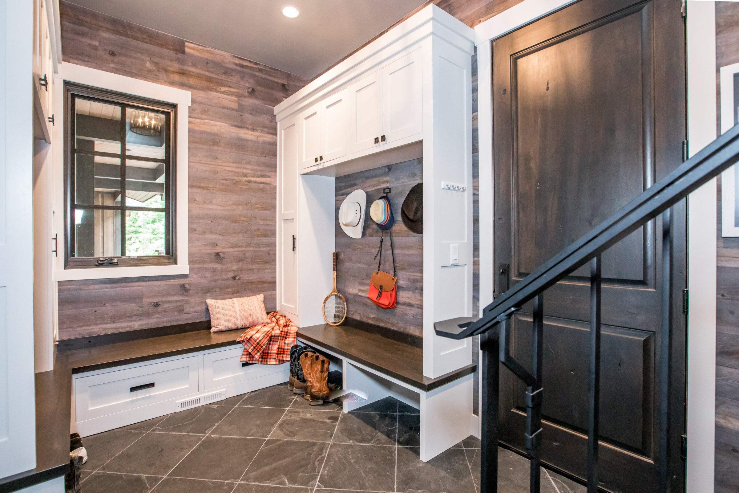 A rustic entryway features a built-in bench and storage, wooden walls, slate flooring, hats hanging, and a staircase with a black handrail.