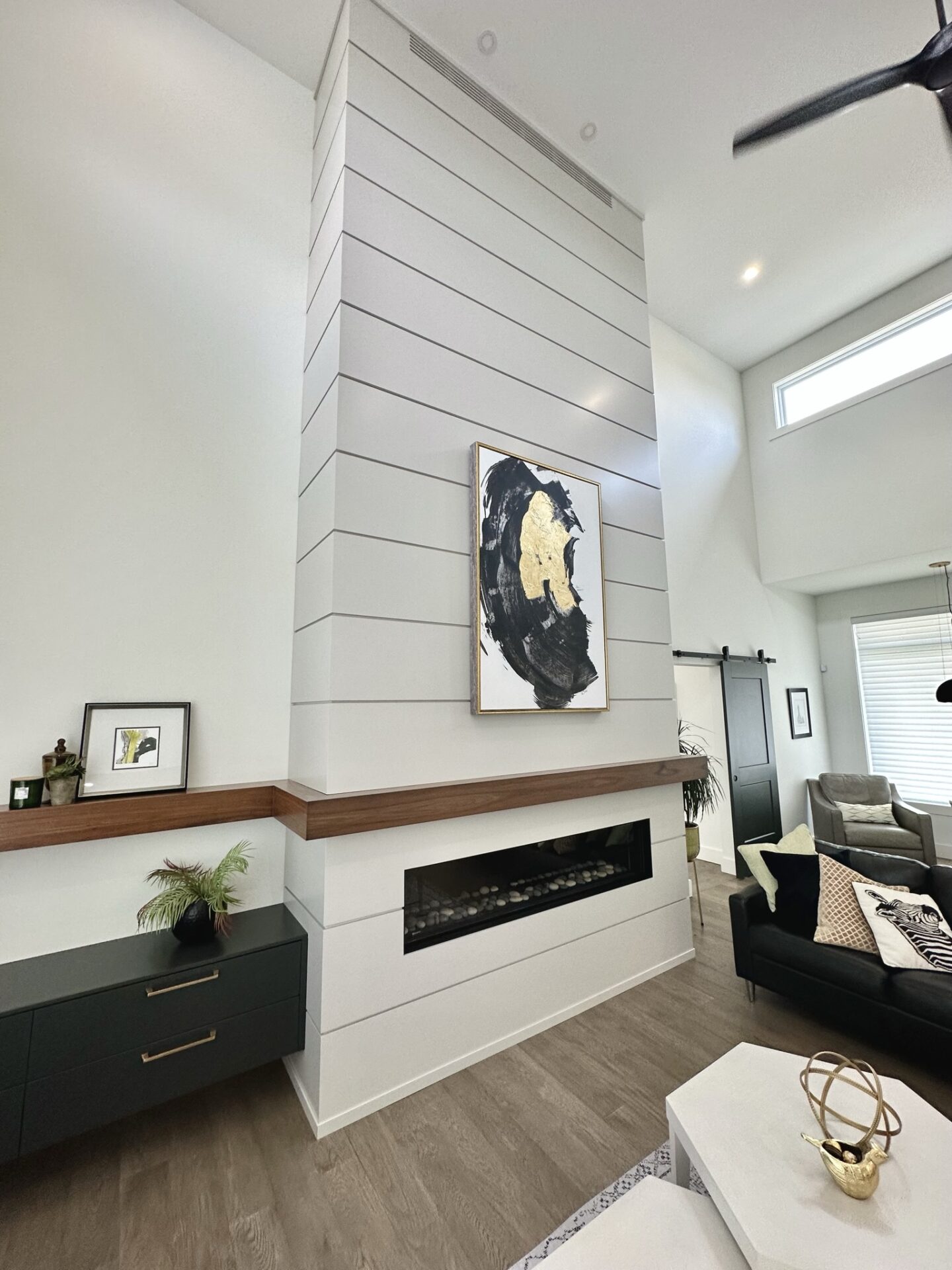 A modern living room featuring a sleek fireplace, shiplap wall with abstract art, dark furniture, decorative items, and an elegant ceiling fan.