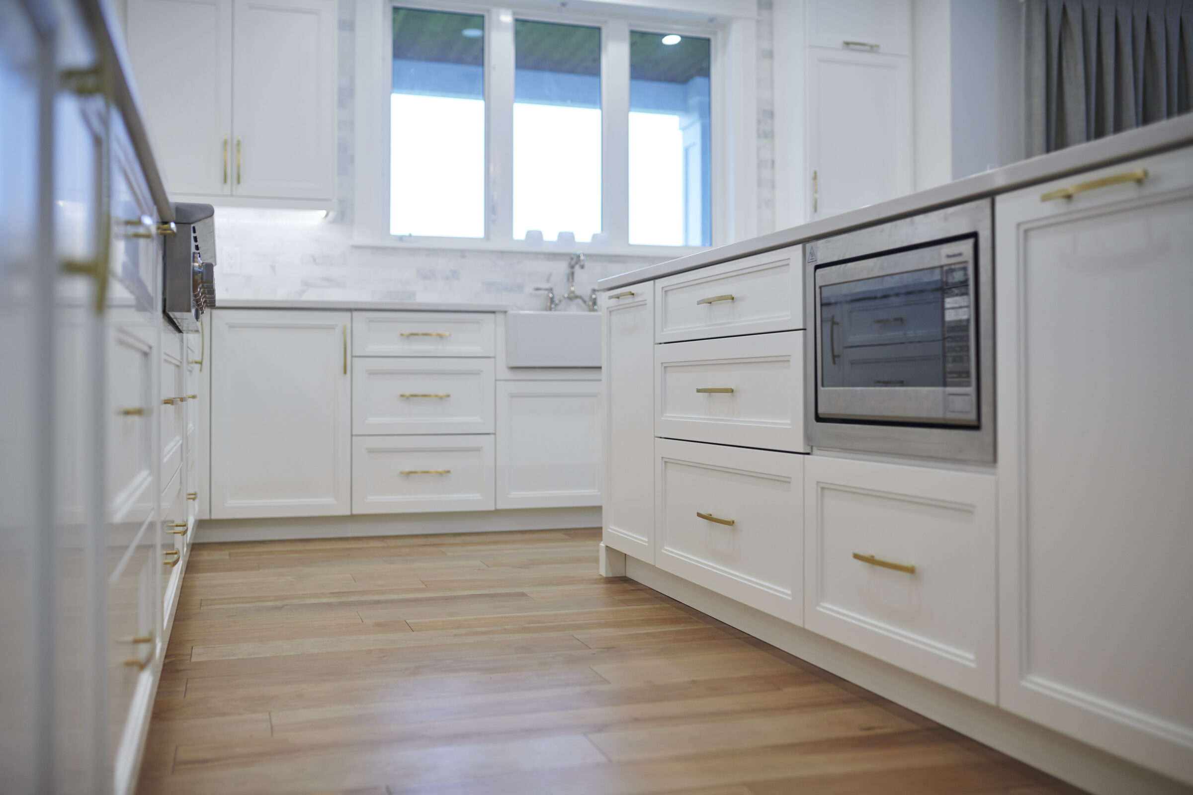 Modern kitchen with white cabinetry, gold handles, built-in oven, marble backsplash, herringbone wooden floor, and windows providing natural light.