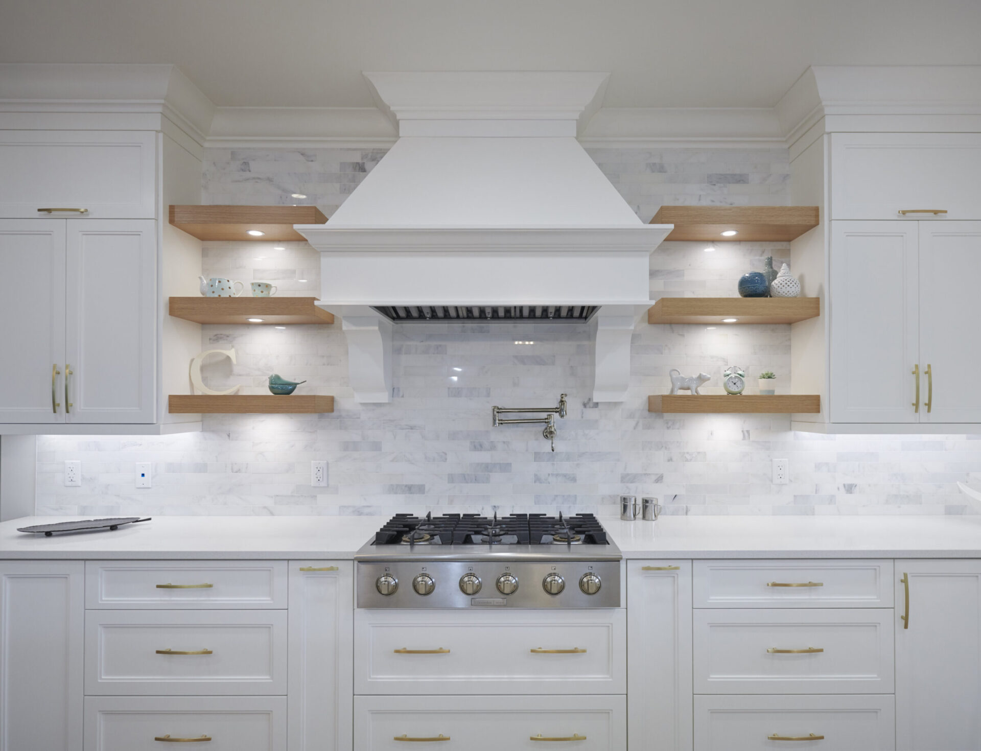 A modern kitchen with white cabinets, marble backsplash, floating wooden shelves, and stainless steel appliances including a gas stove and range hood.