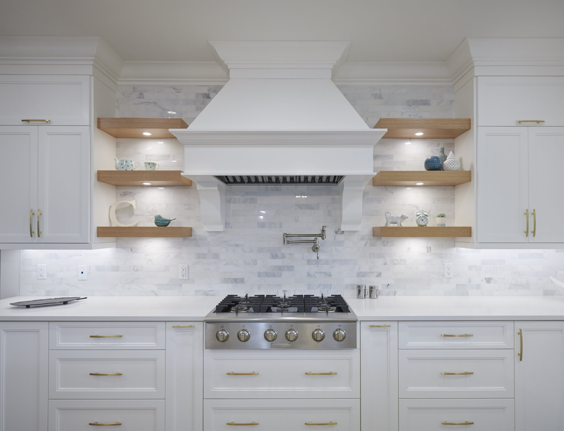 A modern kitchen with white cabinets, marble backsplash, floating wooden shelves, and stainless steel appliances including a gas stove and range hood.