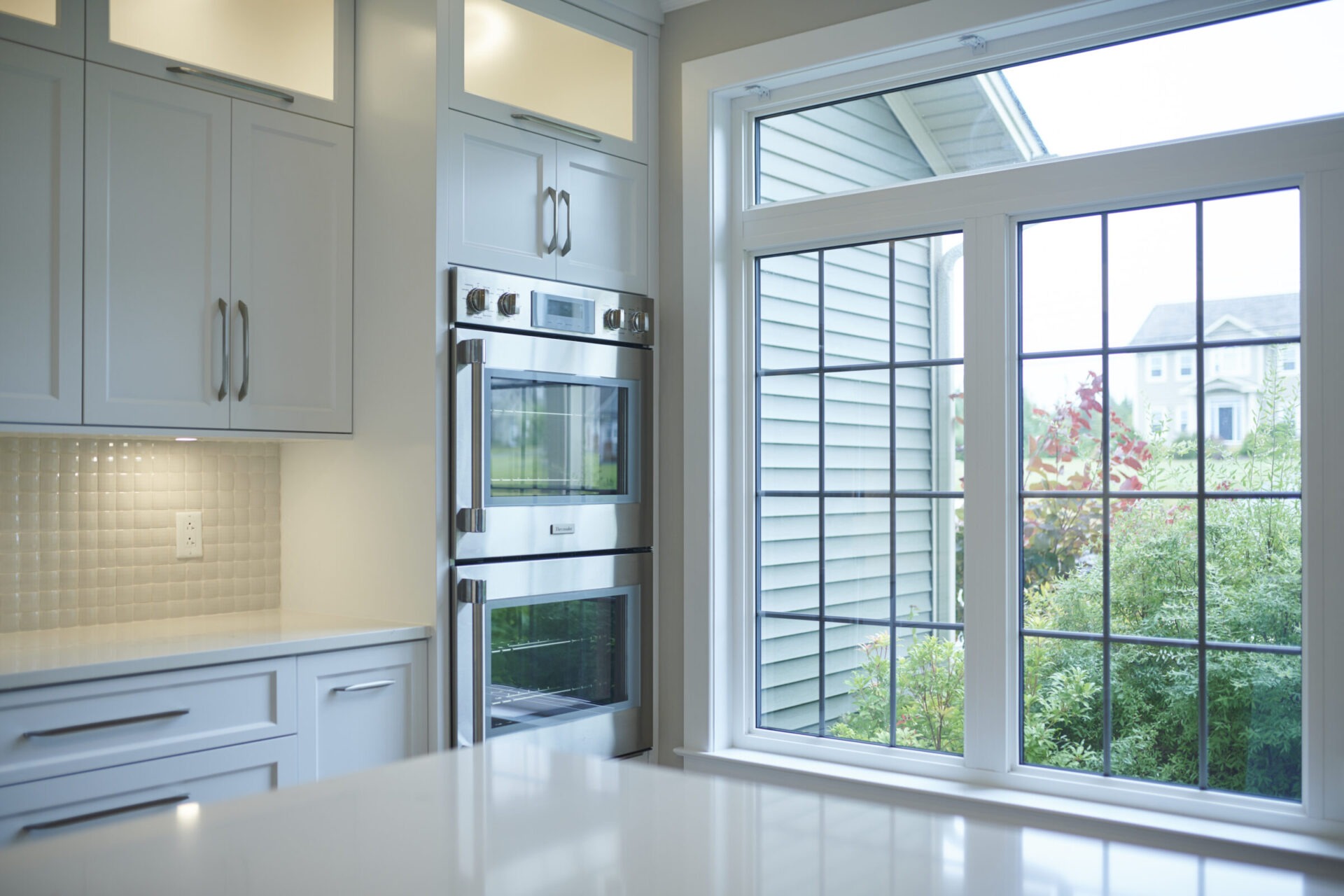 A modern kitchen with white cabinetry, double built-in ovens, and a window offering a view of greenery and neighboring houses. Bright, clean, domestic interior.