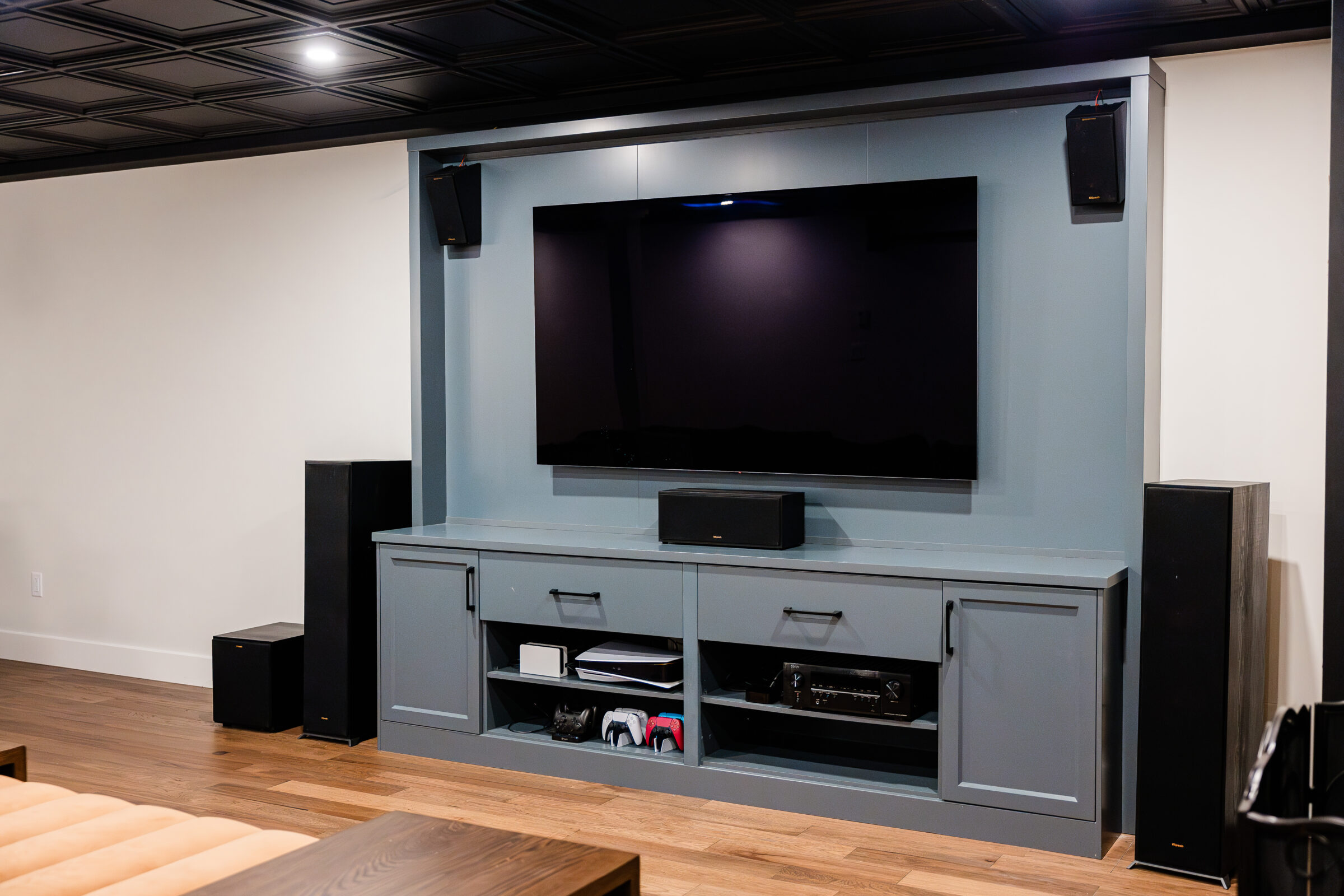 Modern entertainment center with large flat-screen TV, soundbar, two floor-standing speakers, shelving with components, and ceiling-mounted surround speakers in a room.